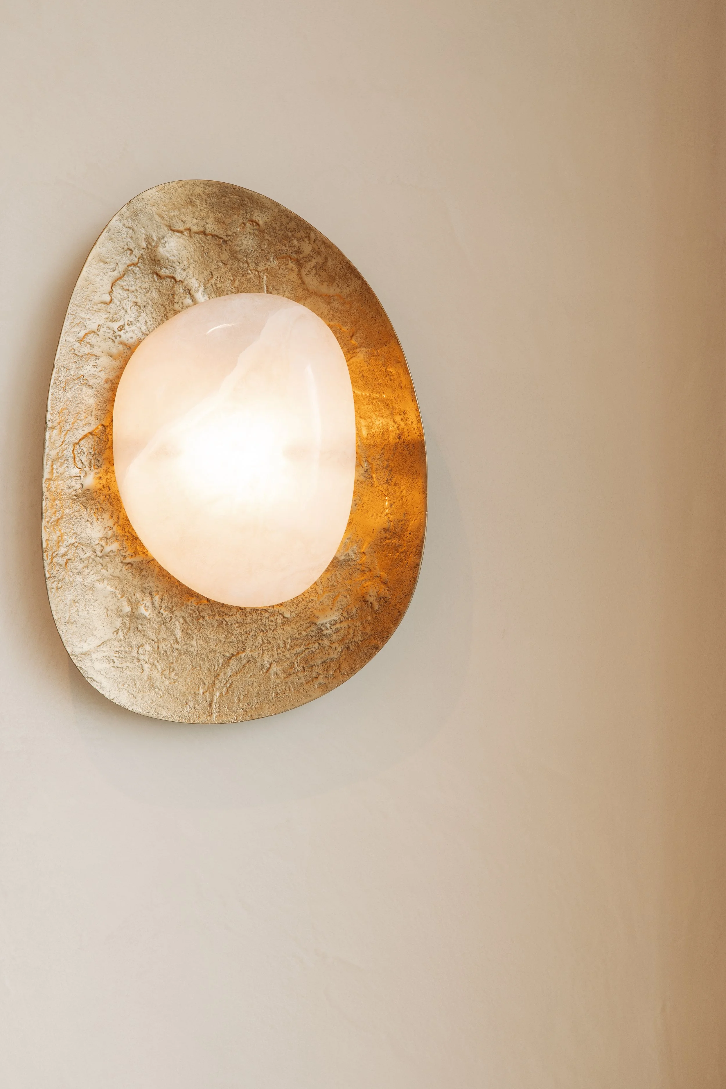 A round light fixture with a white, semi-opaque cover mounted on a decorative gold and textured oval wall plate on a beige wall.