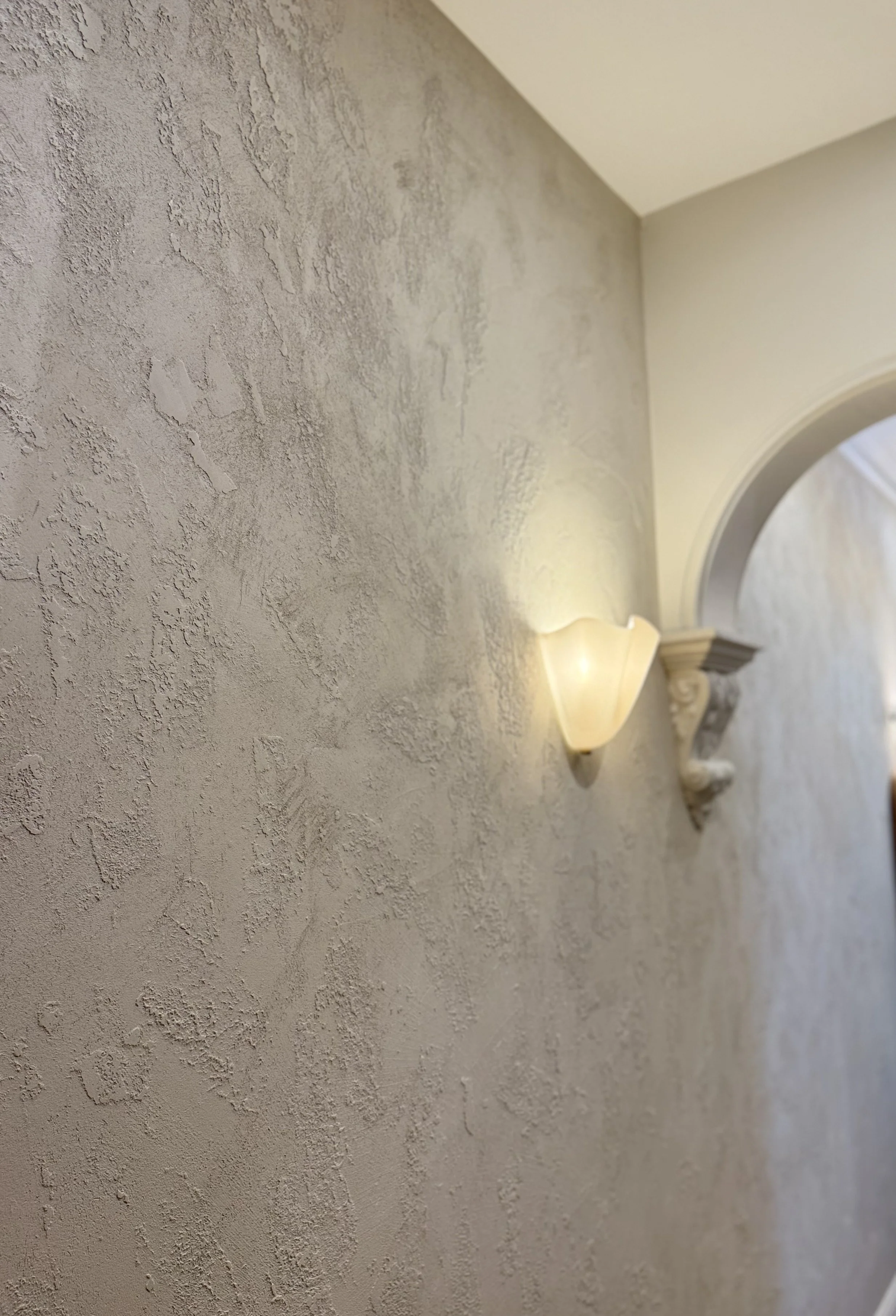 Close-up view of a textured beige wall with a decorative wall sconce illuminating the space and a partial view of an archway at the edge of the image.