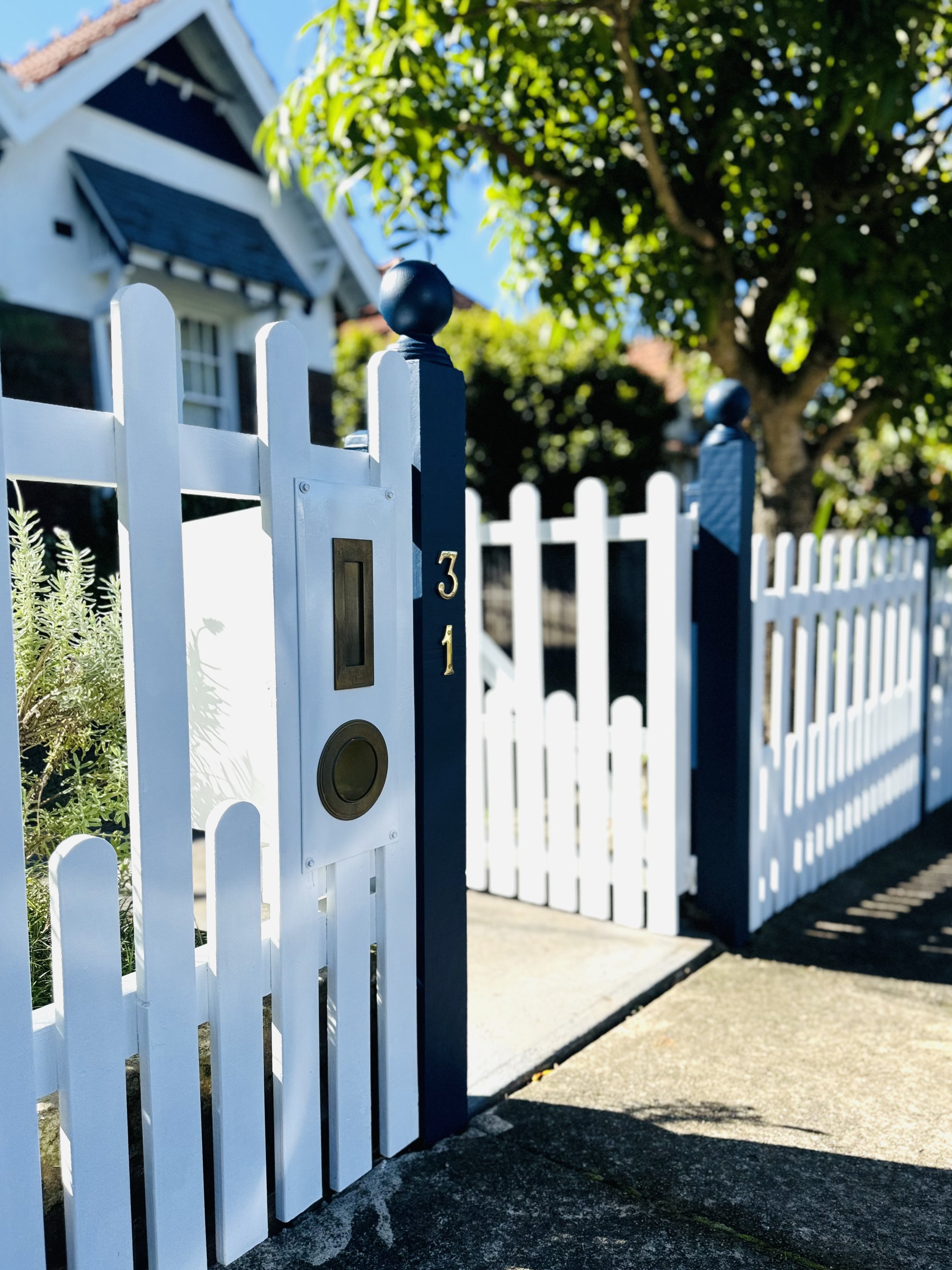 White picket fence with a blue gate and house in background, sunny day with trees and sky.