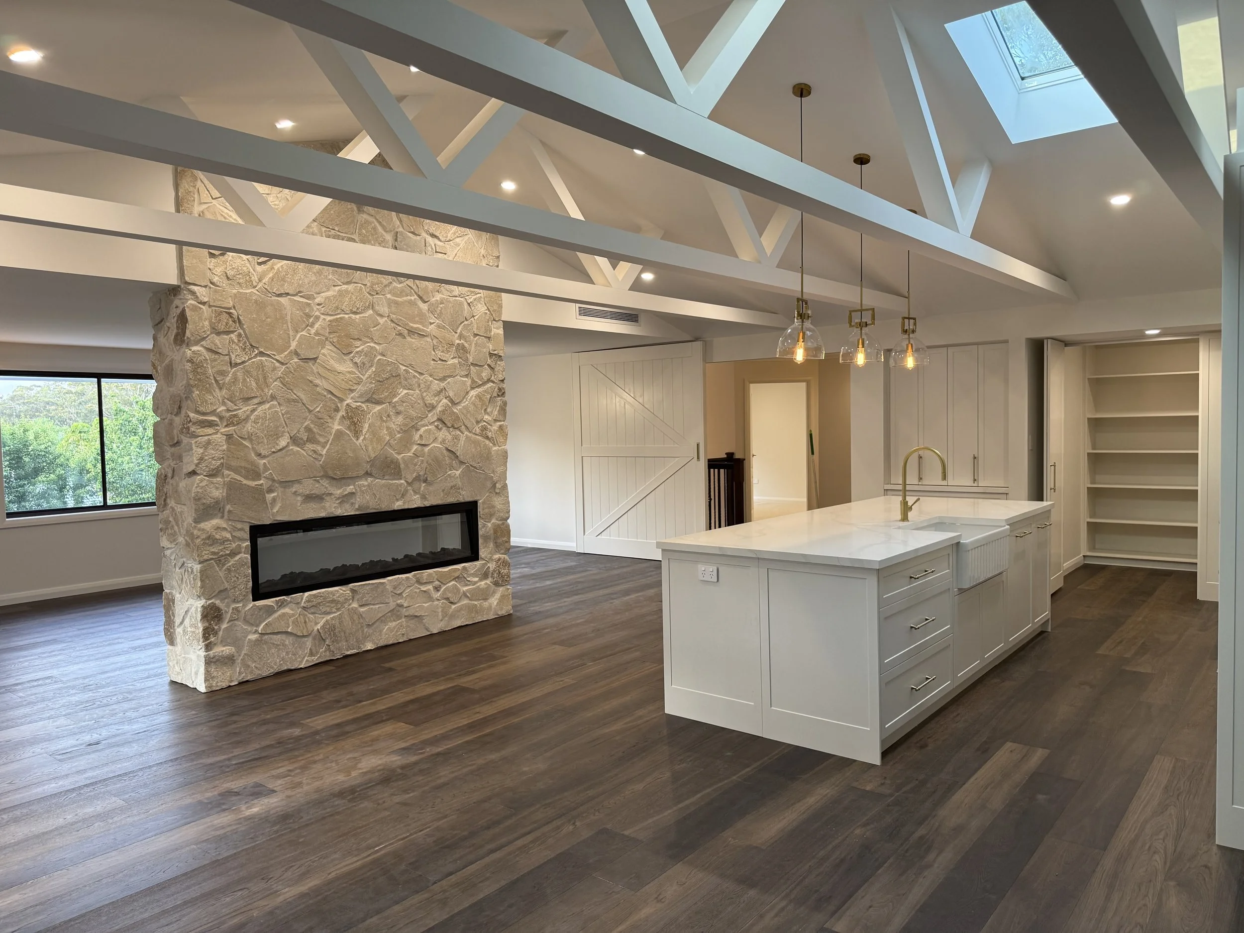 Modern kitchen with white cabinets and a large island, stone accent wall with a fireplace, wood flooring, ceiling with skylights, pendant lights, and a view of greenery outside.
