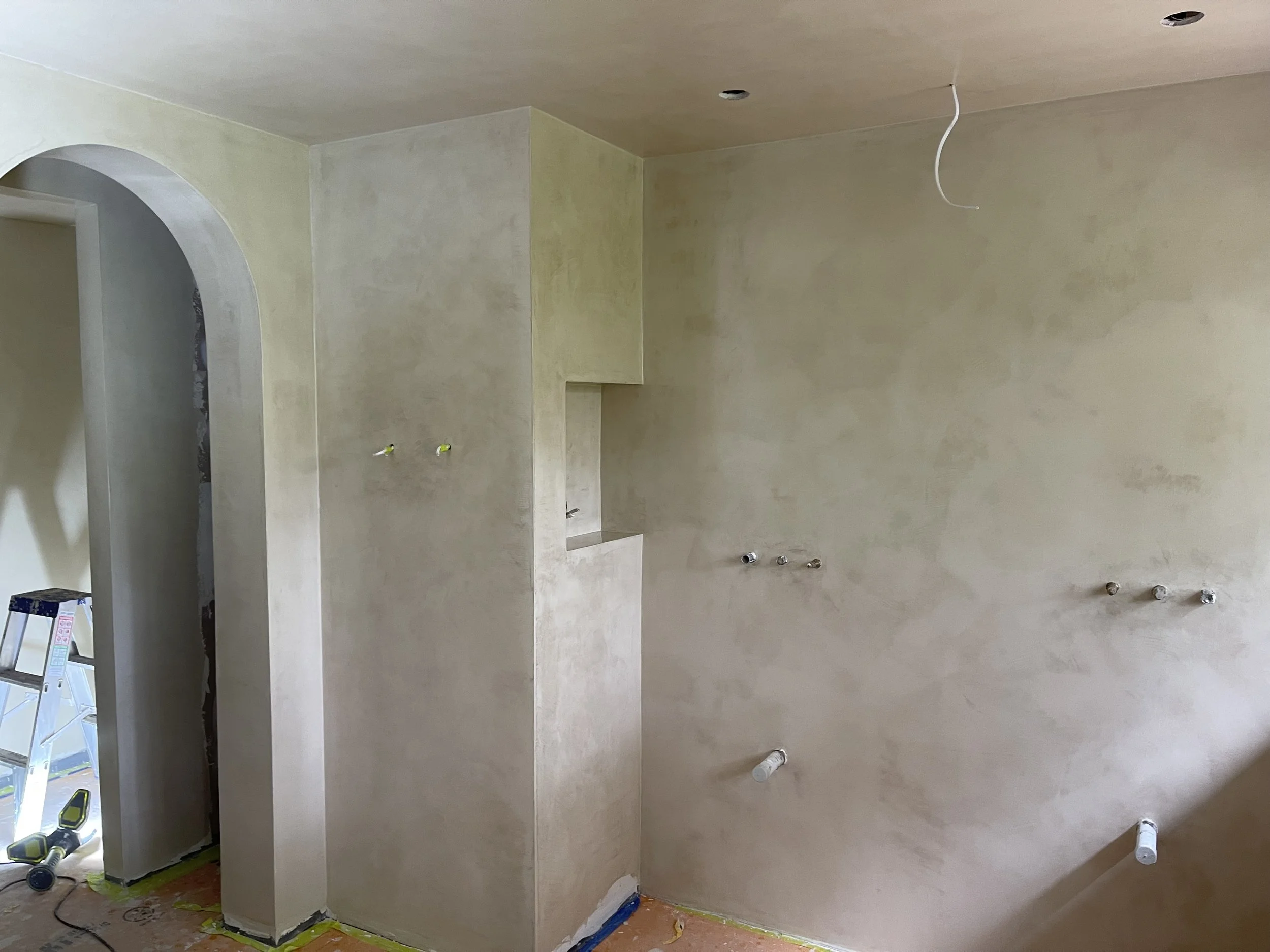 Interior room under construction with drywall, electrical wiring, and installation of fixtures. Tools and construction materials are visible on the floor.