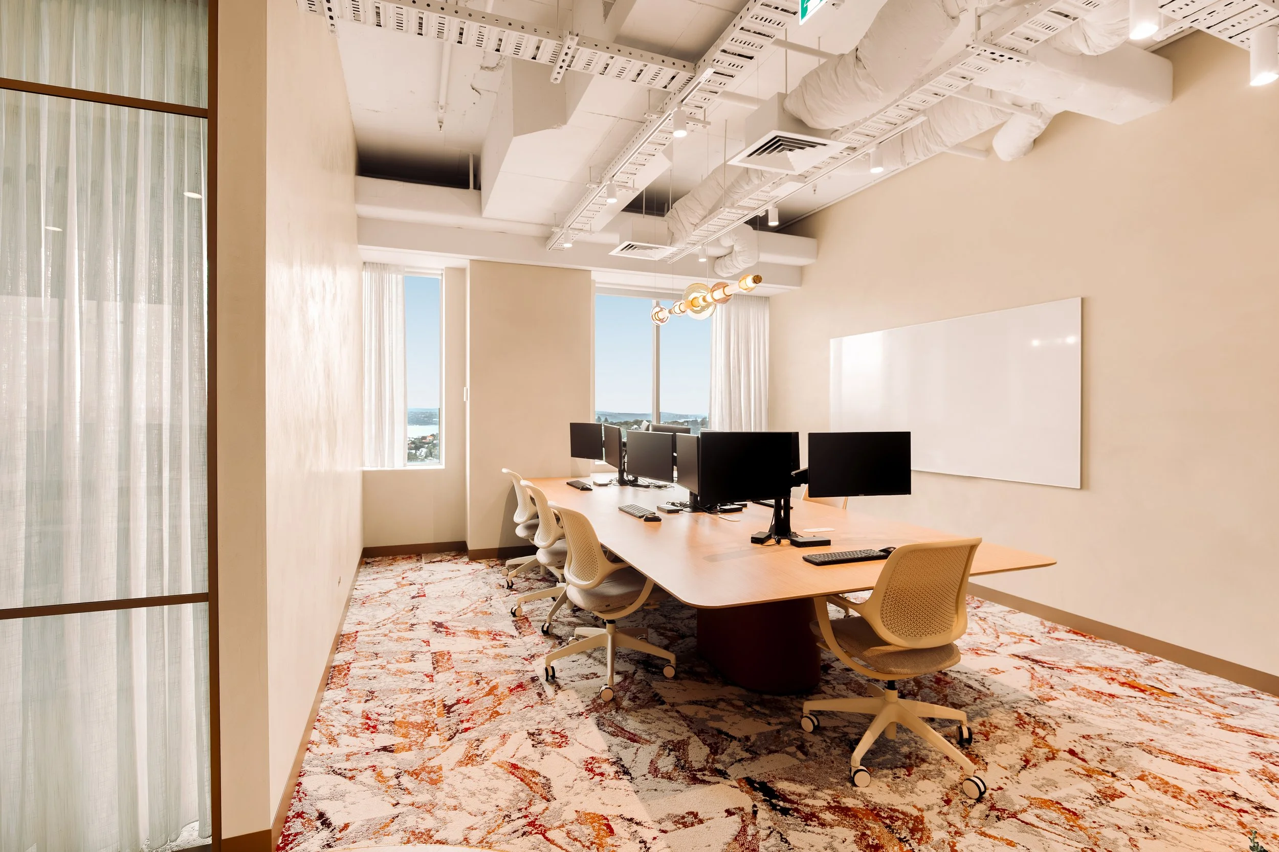 An office conference room with a long wooden table, several black computer monitors, and beige mesh office chairs. The room has large windows with sheer curtains, a colorful patterned rug, and a whiteboard on the wall. The ceiling features exposed wh