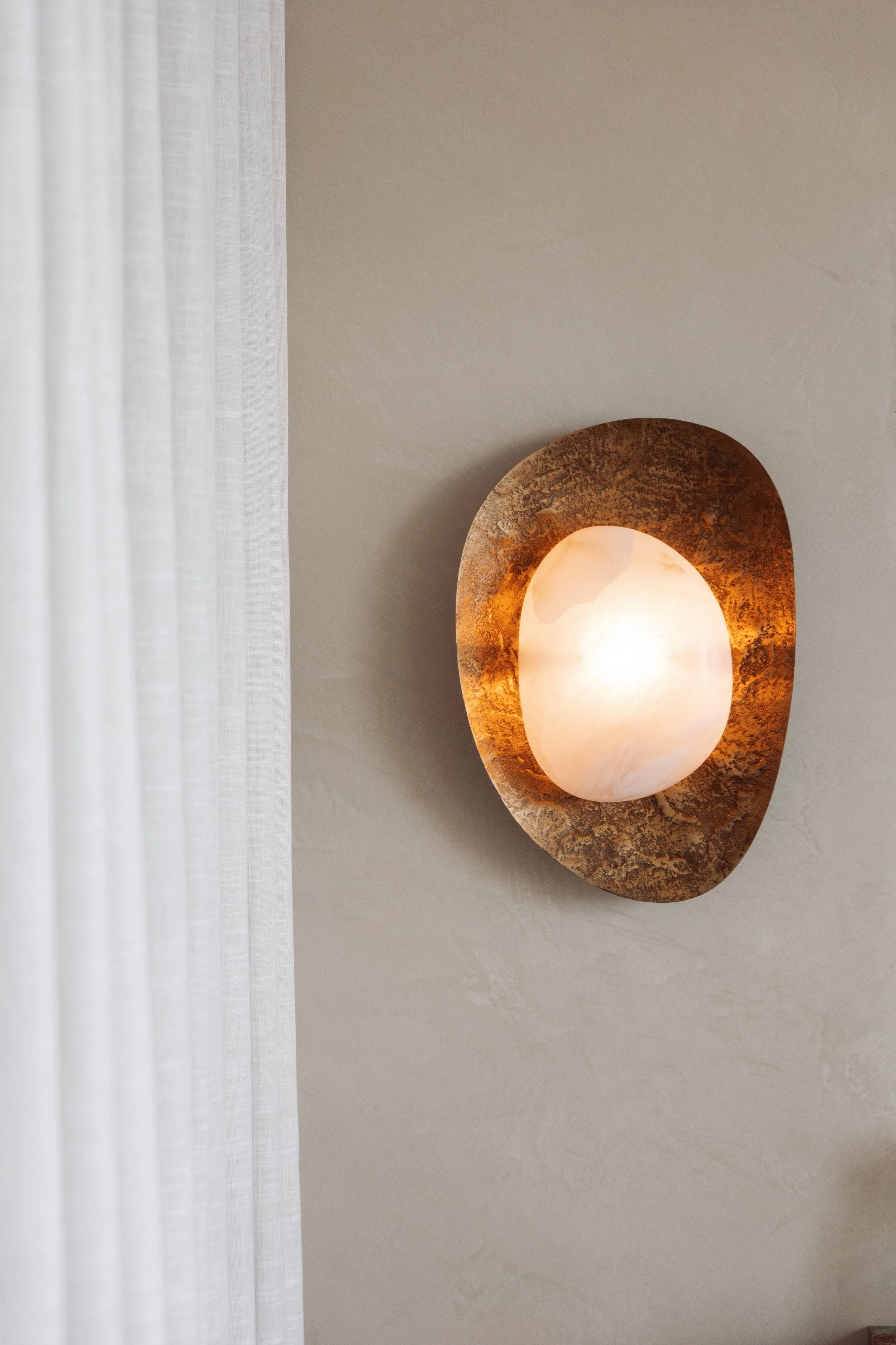Wall-mounted light fixture with a warm glowing light, bordered by a textured bronze or copper oval mount, next to sheer white curtains on a plain white wall.