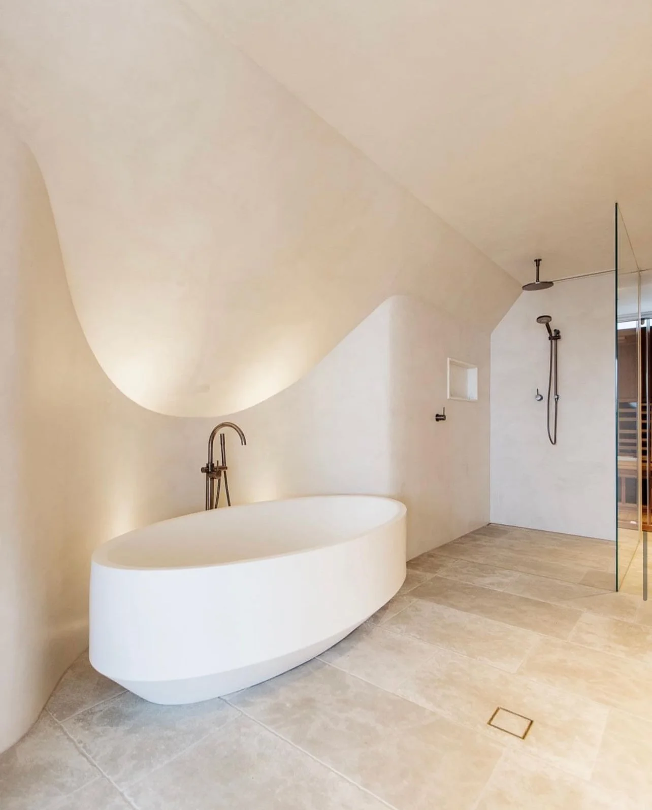 A modern bathroom with a freestanding oval bathtub, a wall-mounted faucet, a walk-in shower with a rain shower head and handheld shower, beige tiled floors, and minimalistic design elements.