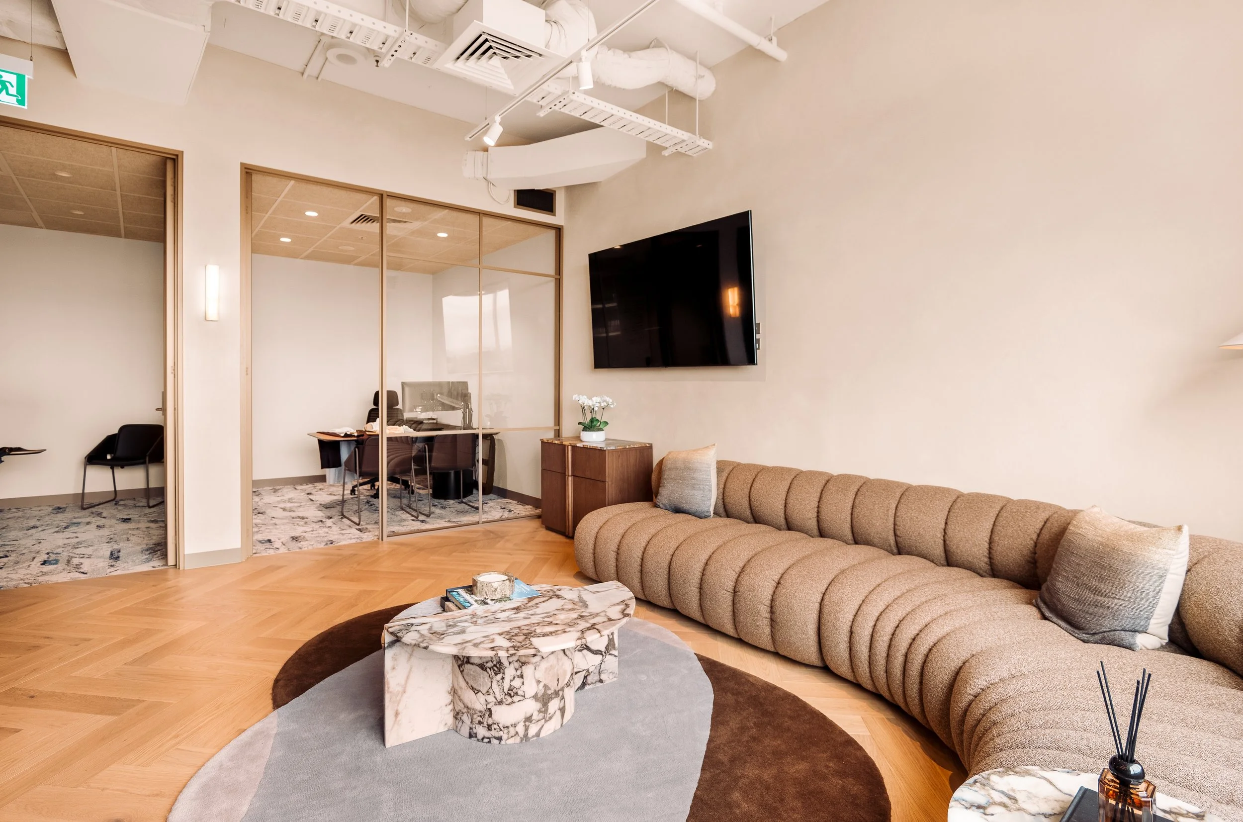Modern office waiting area with beige curved sofa, marble coffee table, and large wall-mounted TV; glass-walled office with desk and chair in the background.