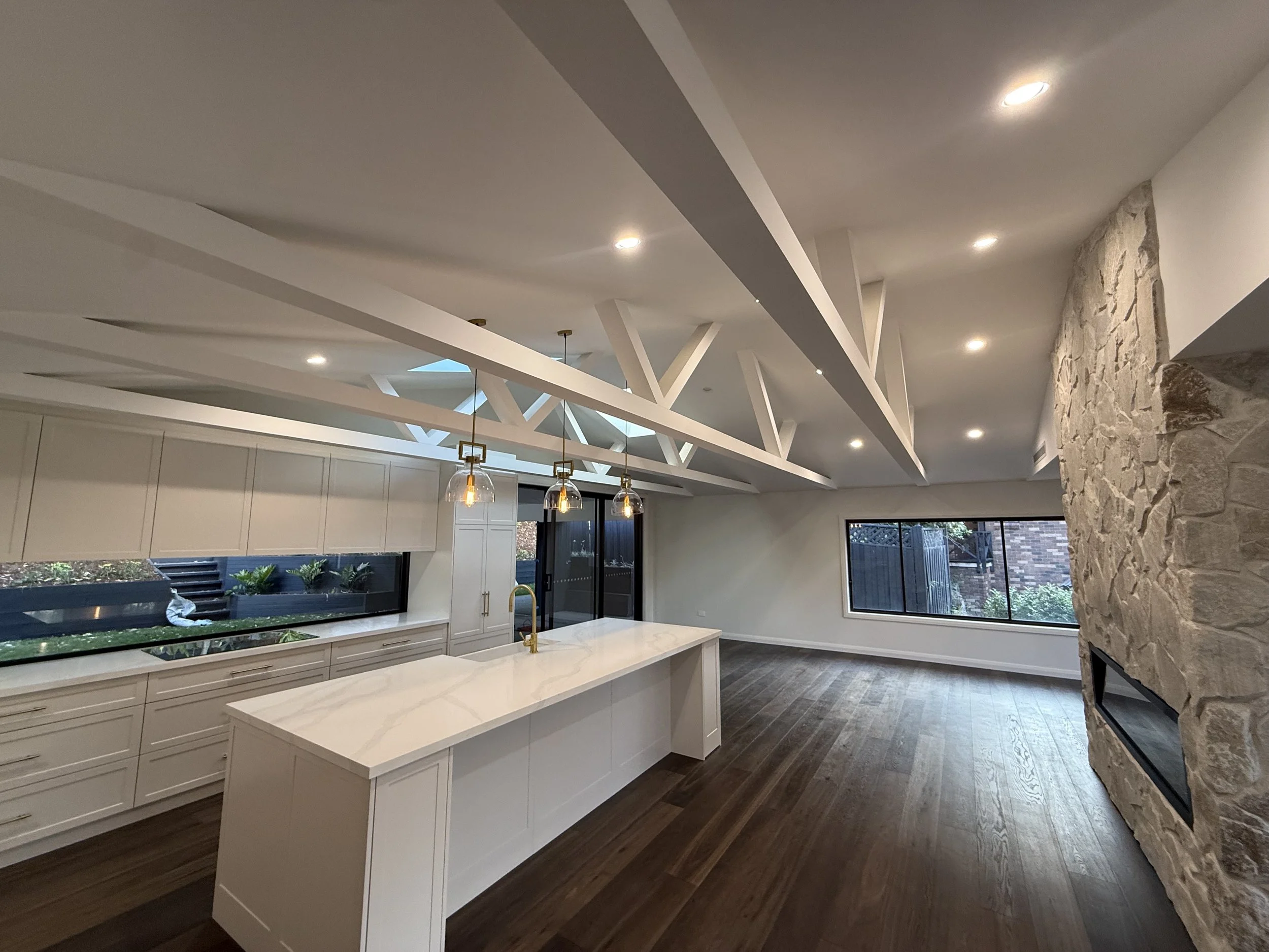 Modern kitchen with white cabinetry, marble island, gold accents, pendant lighting, large windows, and a stone feature wall.