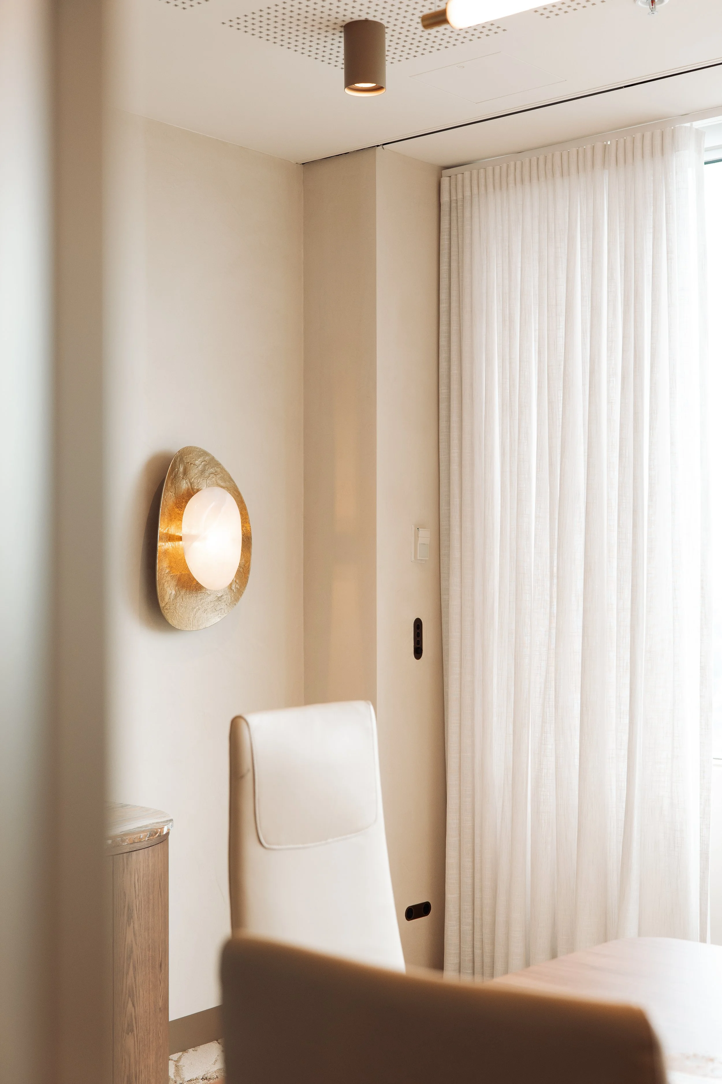 A corner of a room with a beige curtain, a white chair, a wall-mounted light fixture with a round, gold-colored frame and a white globe, and a beige wall with electrical outlets. There is also a small wooden side table with a marble top.