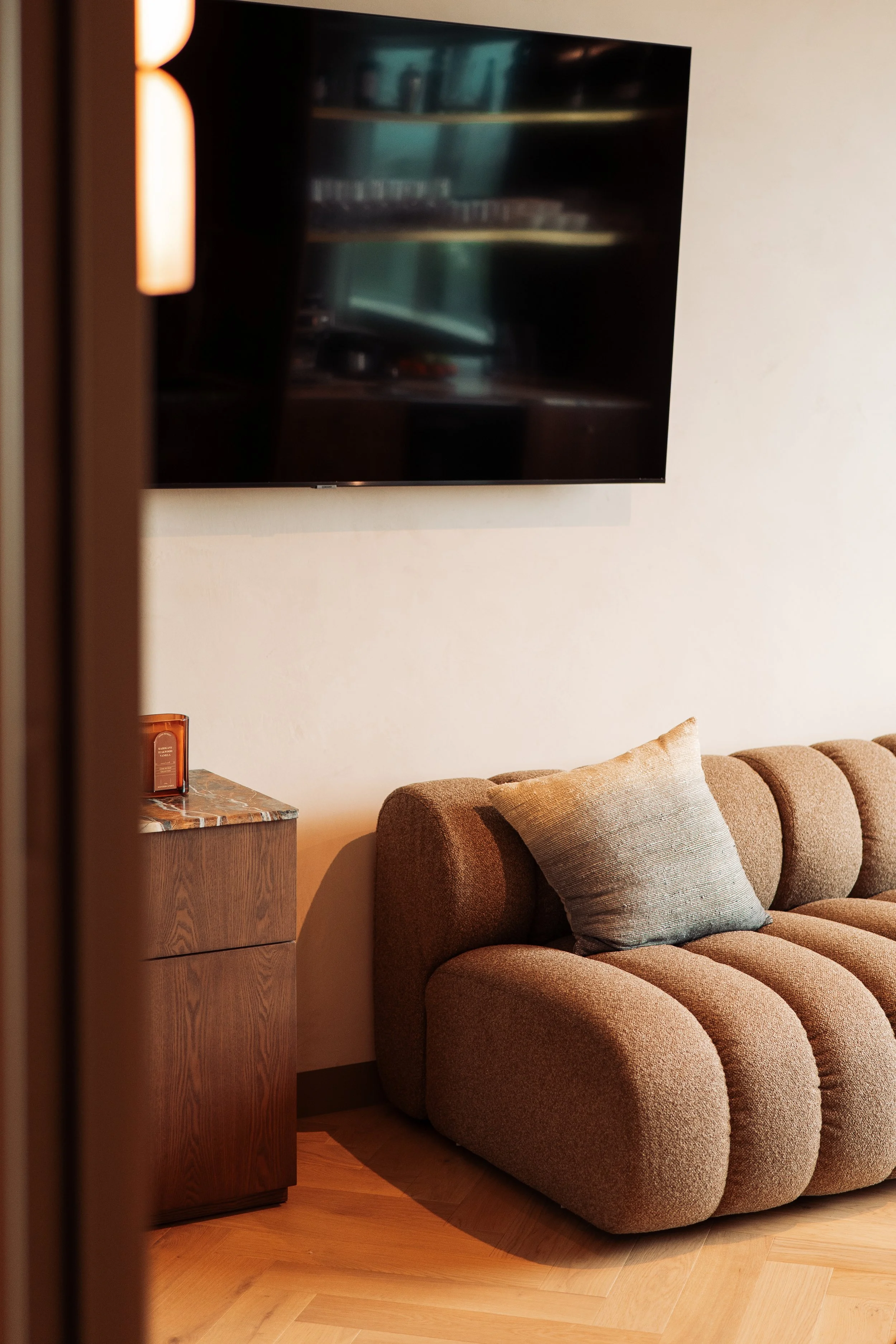 A living room area with a brown upholstered couch, a beige pillow, a wooden side table, a wall-mounted flat-screen TV, and a corner of a wall with a light color.