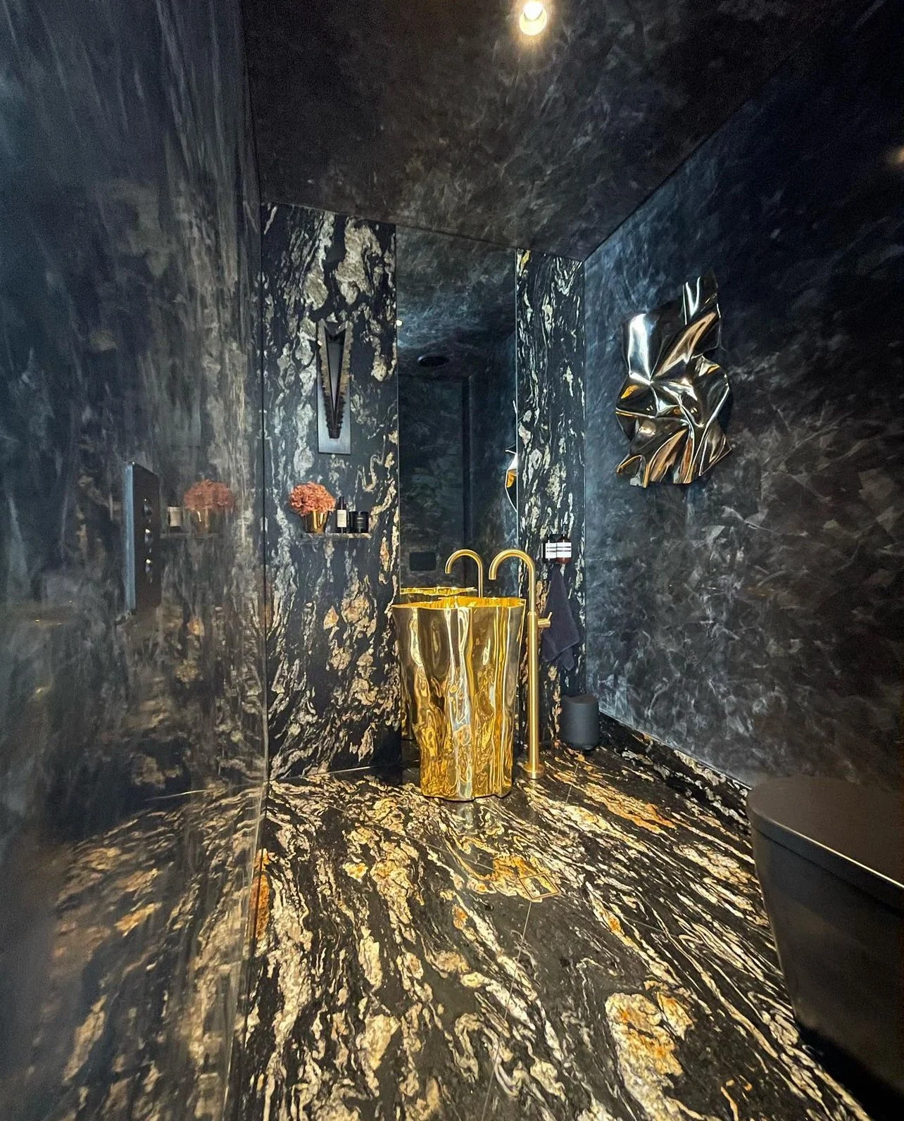 Luxurious bathroom with black marble walls and floor with gold veining, featuring a gold-colored freestanding bathtub with matching faucet, decorative wall art, small shelves with decor, and a black toilet.