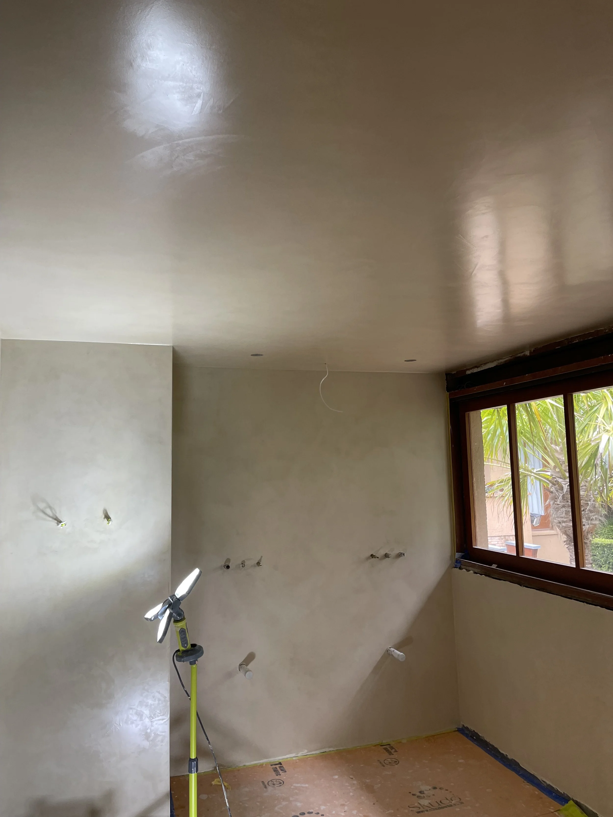 Interior room under renovation with unfinished drywall on the walls, a window with greenery outside, and a work light on a stand illuminating the space.