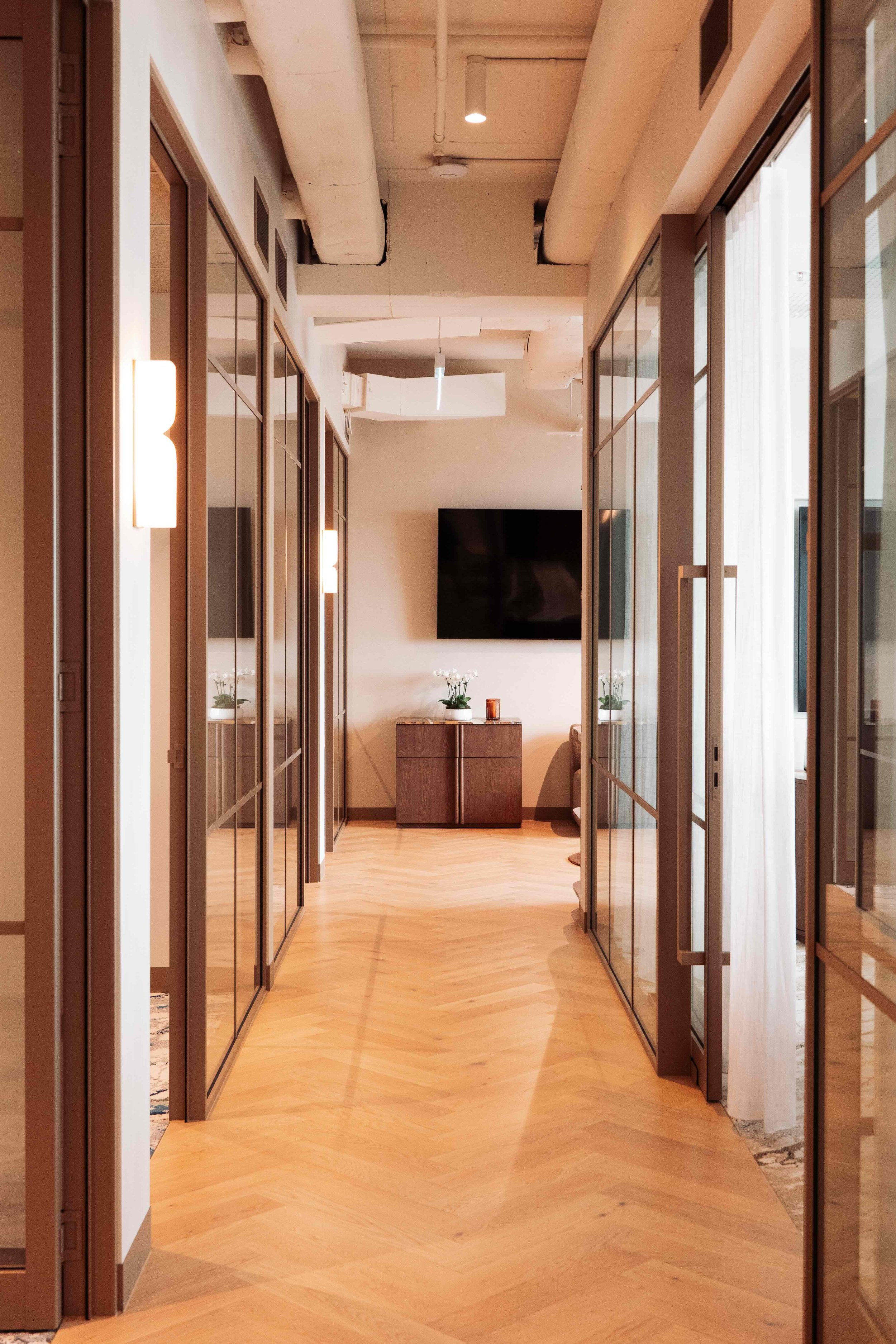 A hallway with wooden flooring and glass-panelled doors on either side, leading to a room with a wall-mounted television and a wooden table with plants and decorative items, illuminated by wall-mounted lights and natural light from large windows.