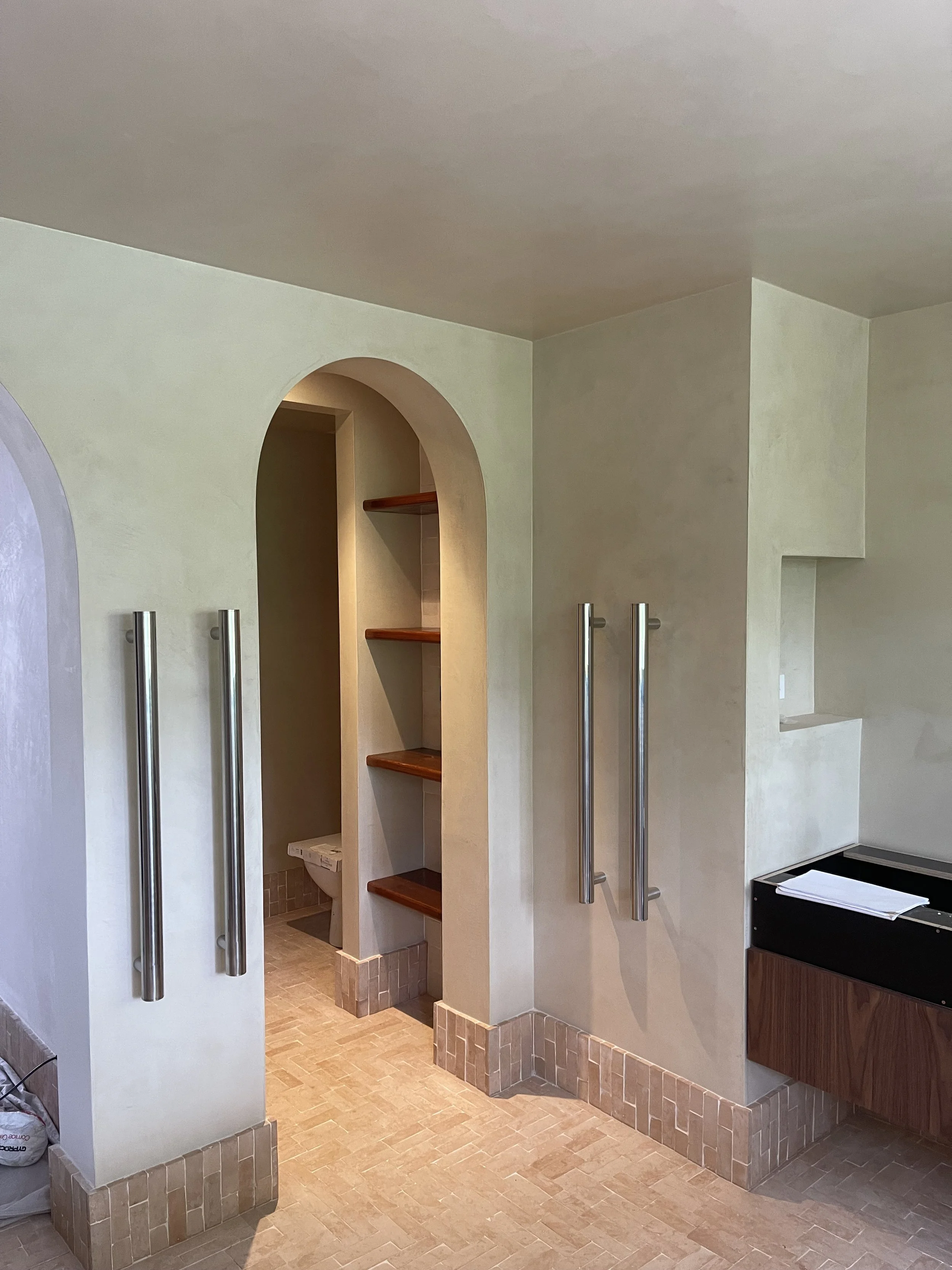 Public restroom with beige walls, arched entrance to toilet stall, wooden shelves inside, and metal grab bars on the walls.