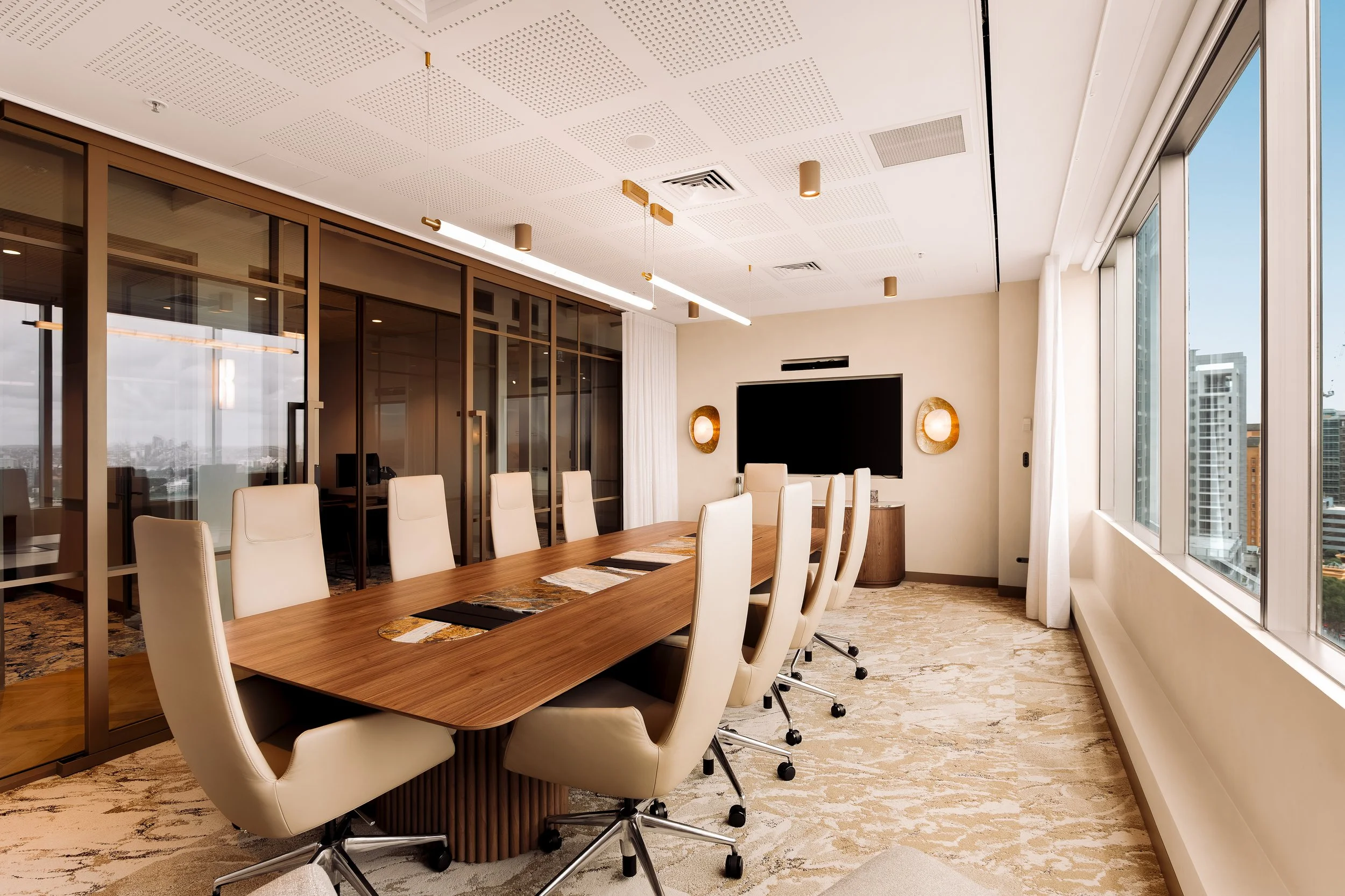 Modern conference room with white chairs around a large wooden table, large windows on the right, and a wall-mounted TV.