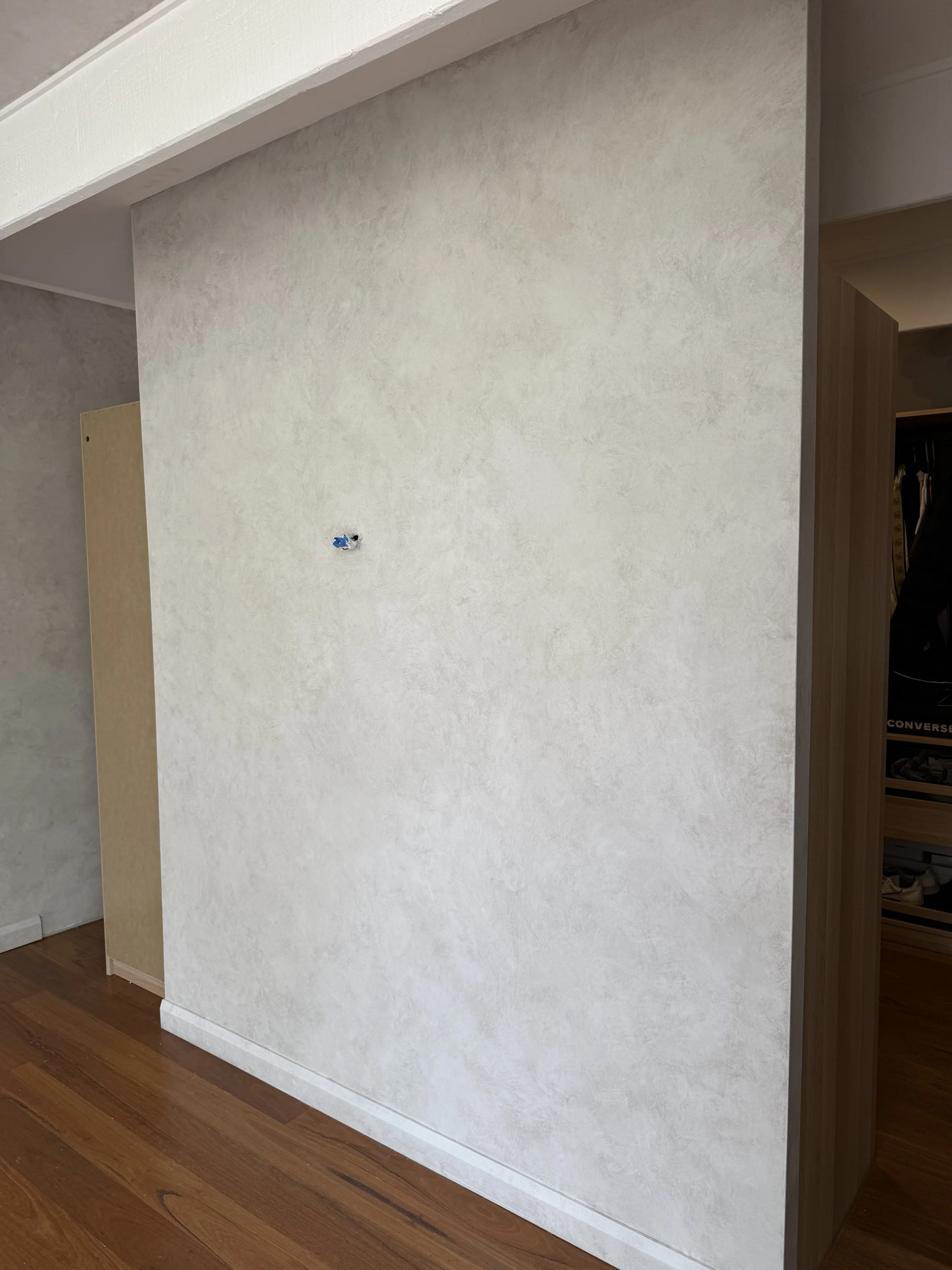 A beige textured wall with an electrical outlet near the center, a corner with another wall and doorway on the right, and a wooden floor.