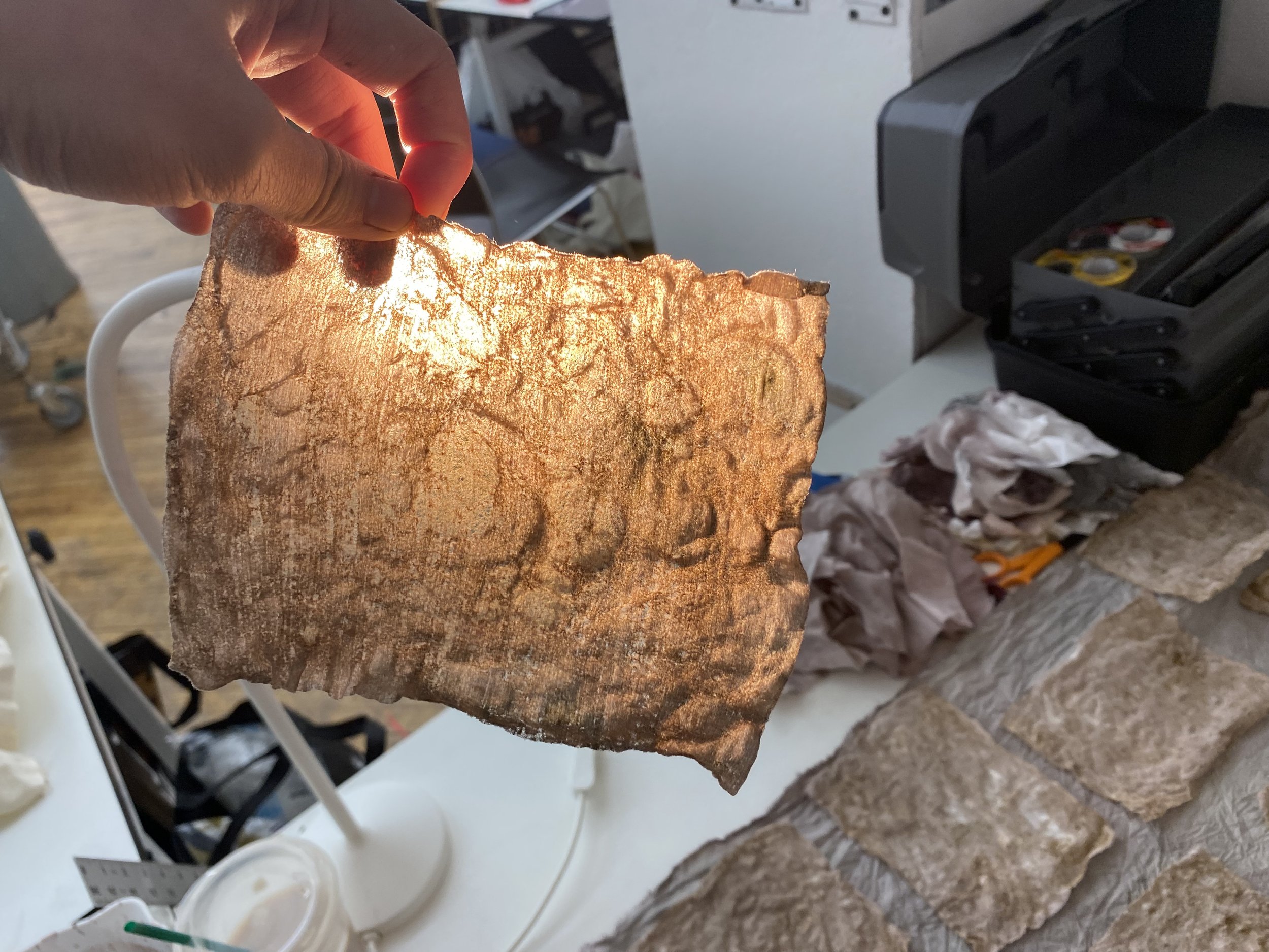 A person's hand holding a piece of fabric with light illuminating it from behind. In the background, a workspace with a printer, crumpled paper, and craft supplies can be seen.