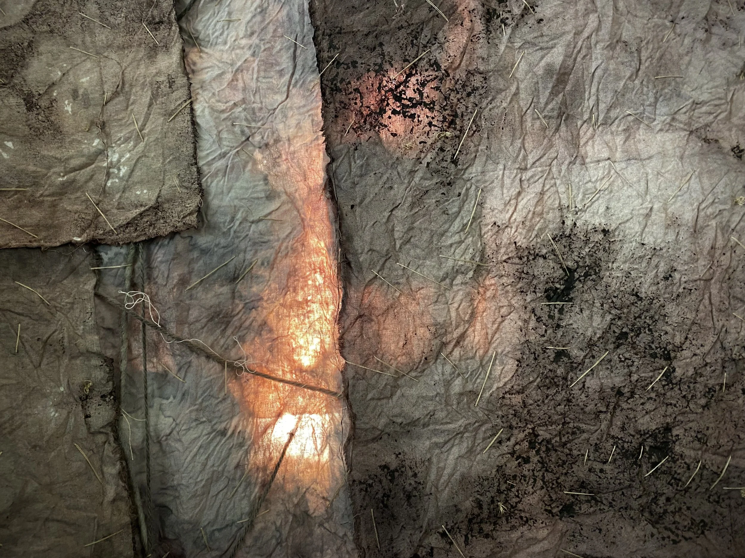 Close-up of a section of fabric or paper with sewn or glued black and red ink or paint splatters, dirt, and small yellowish fibers or threads scattered across the surface.