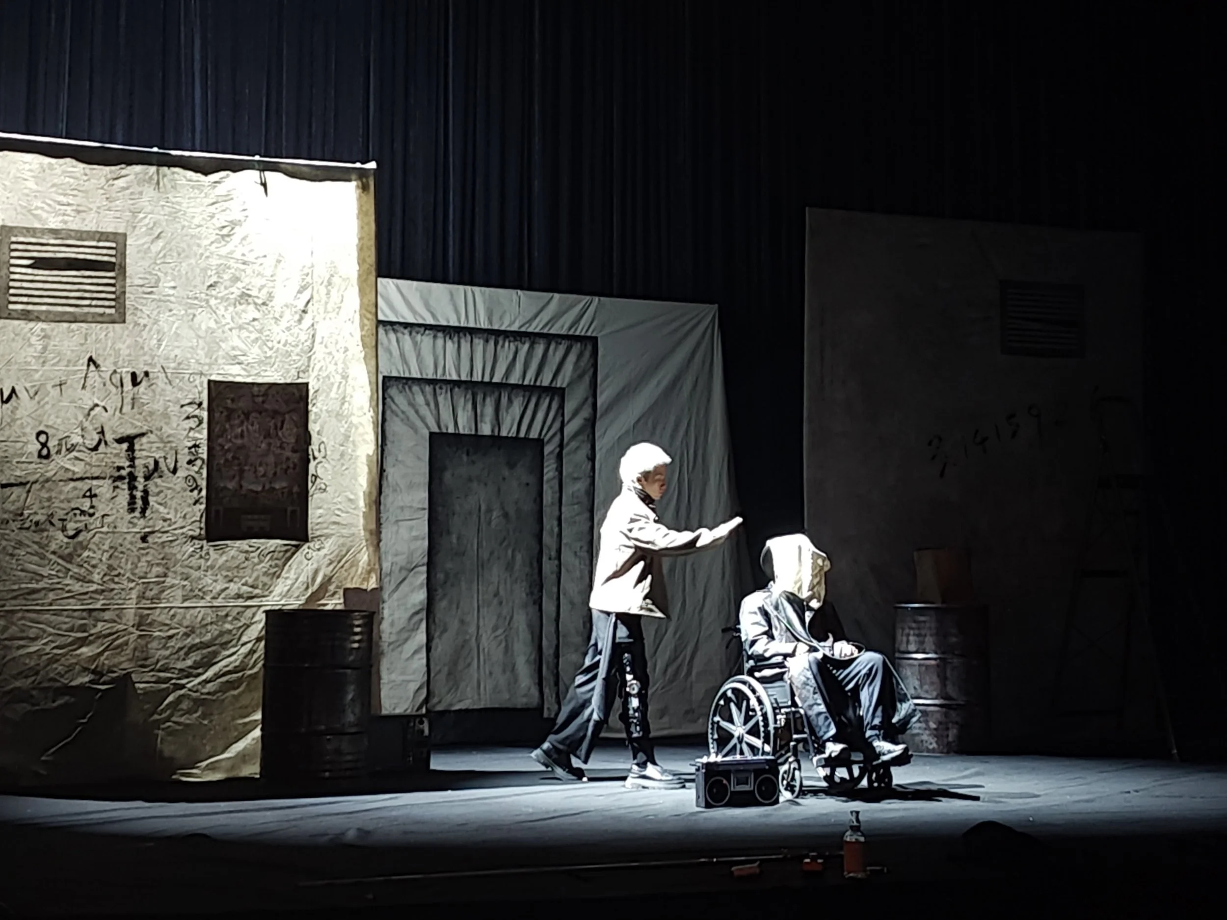 A theatrical stage with a backdrop, featuring a large beige textile with writings and fake structures, and two individuals in costumes, with one in a wheelchair. Various props and equipment are on the stage.