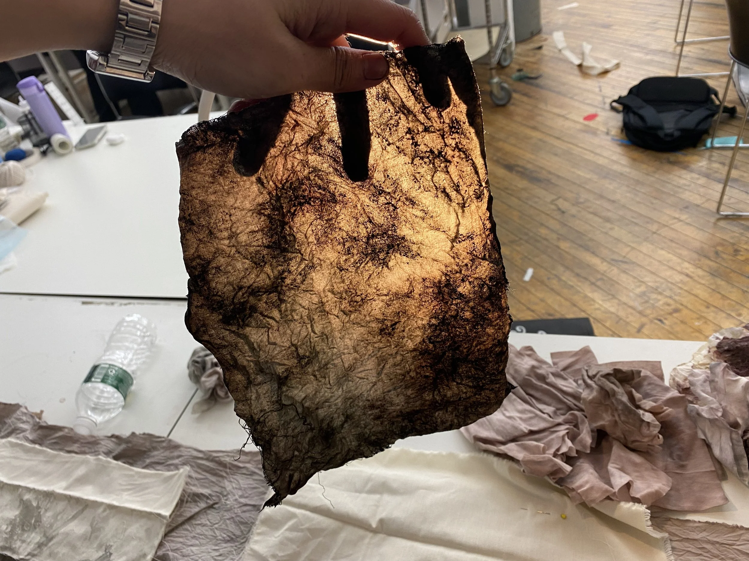 A hand holding a piece of textured, stained, and charred paper with a blackened top edge, in a room with scattered trash, a black bag, a water bottle, and a white table surface.