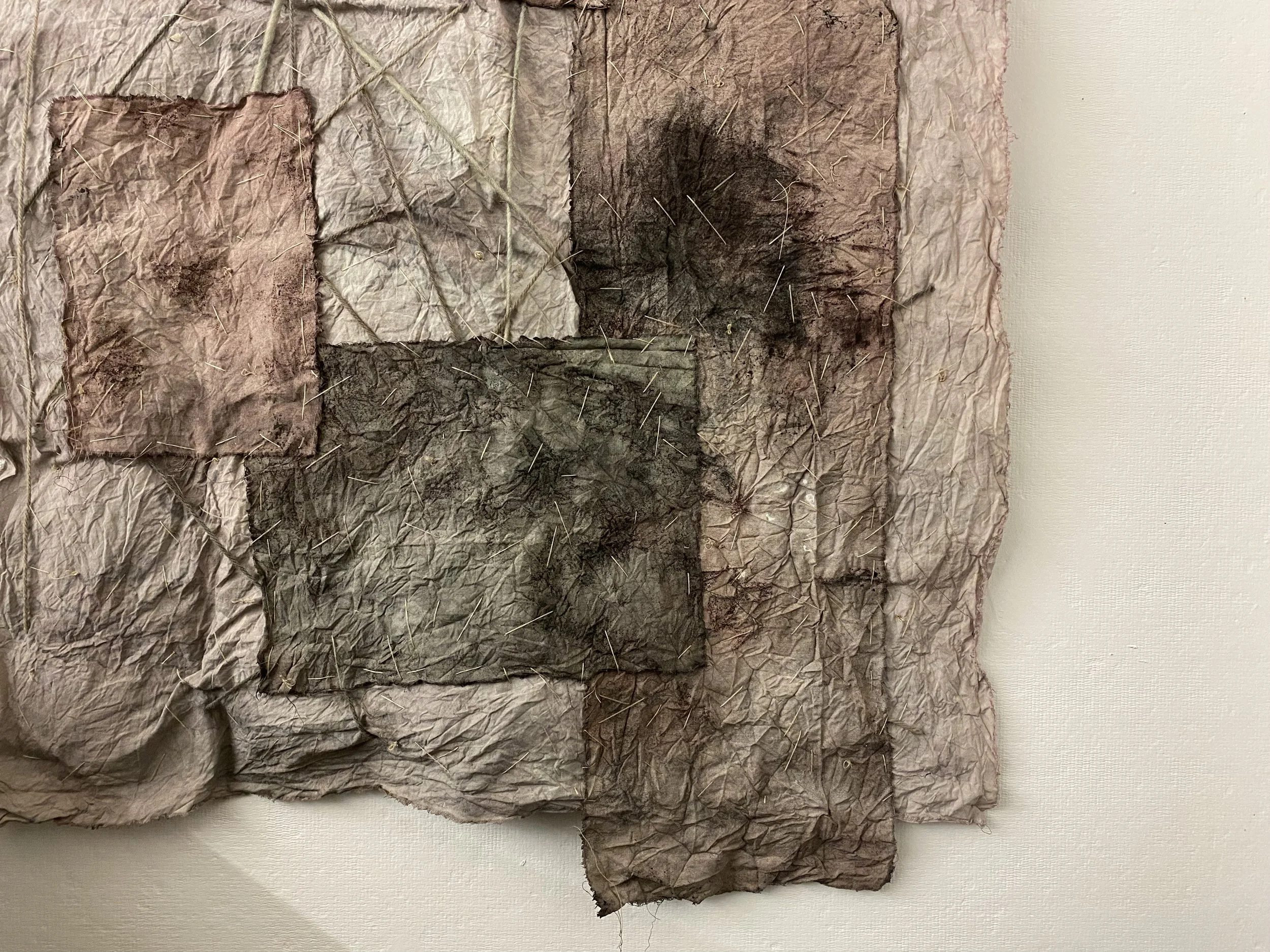 A textured artwork made of crumpled and folded paper, with stitched and painted dark brown and black marks, mounted on a white wall.