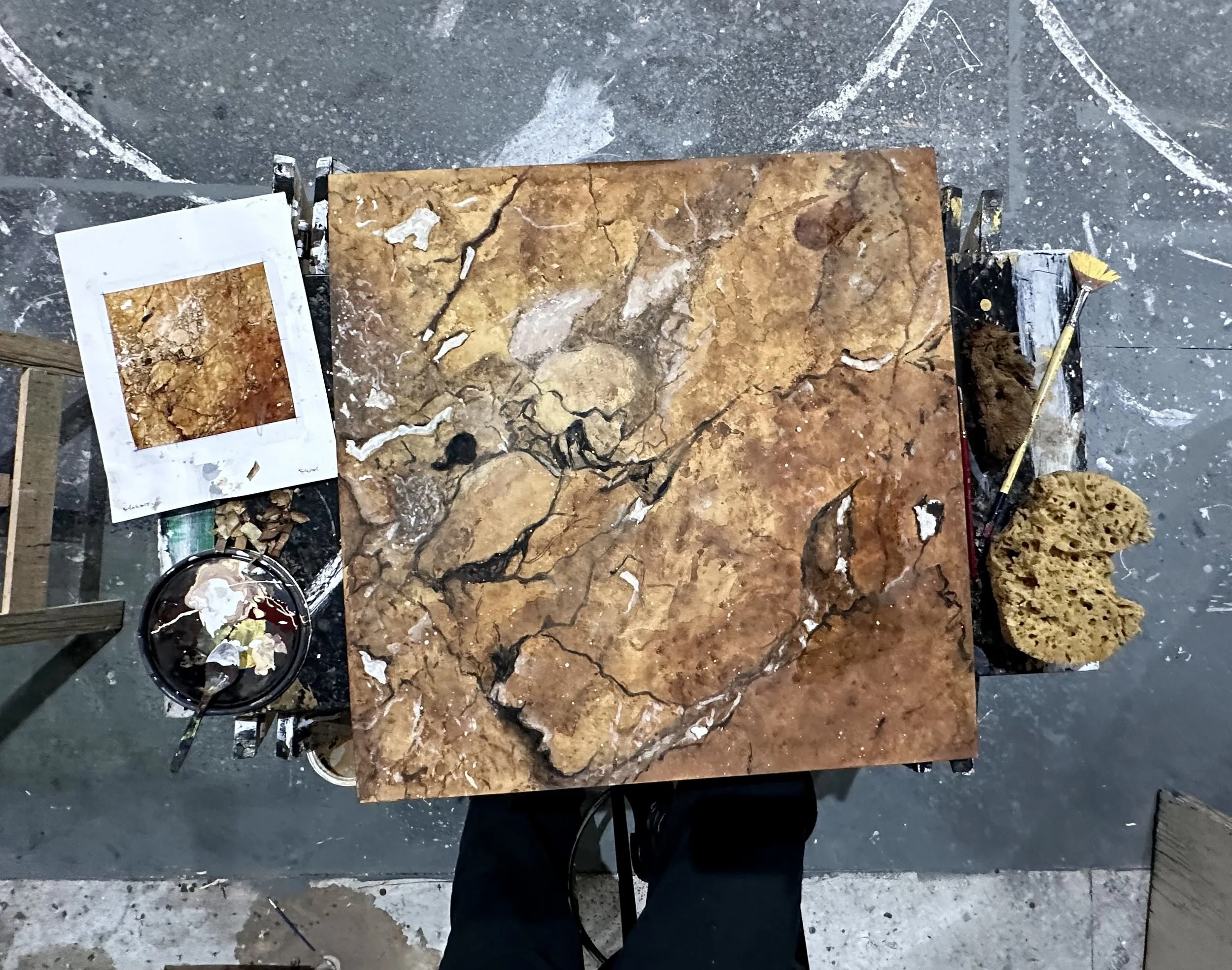 A workspace with a large, polished, orange and brown marble slab on a black table, surrounded by smaller pieces of material, paint supplies, and tools.