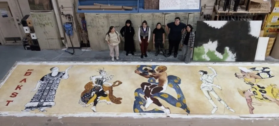 A group of seven people standing above a large, colorful painting on the floor depicting multiple Asian-inspired characters, in what appears to be a warehouse or art studio.