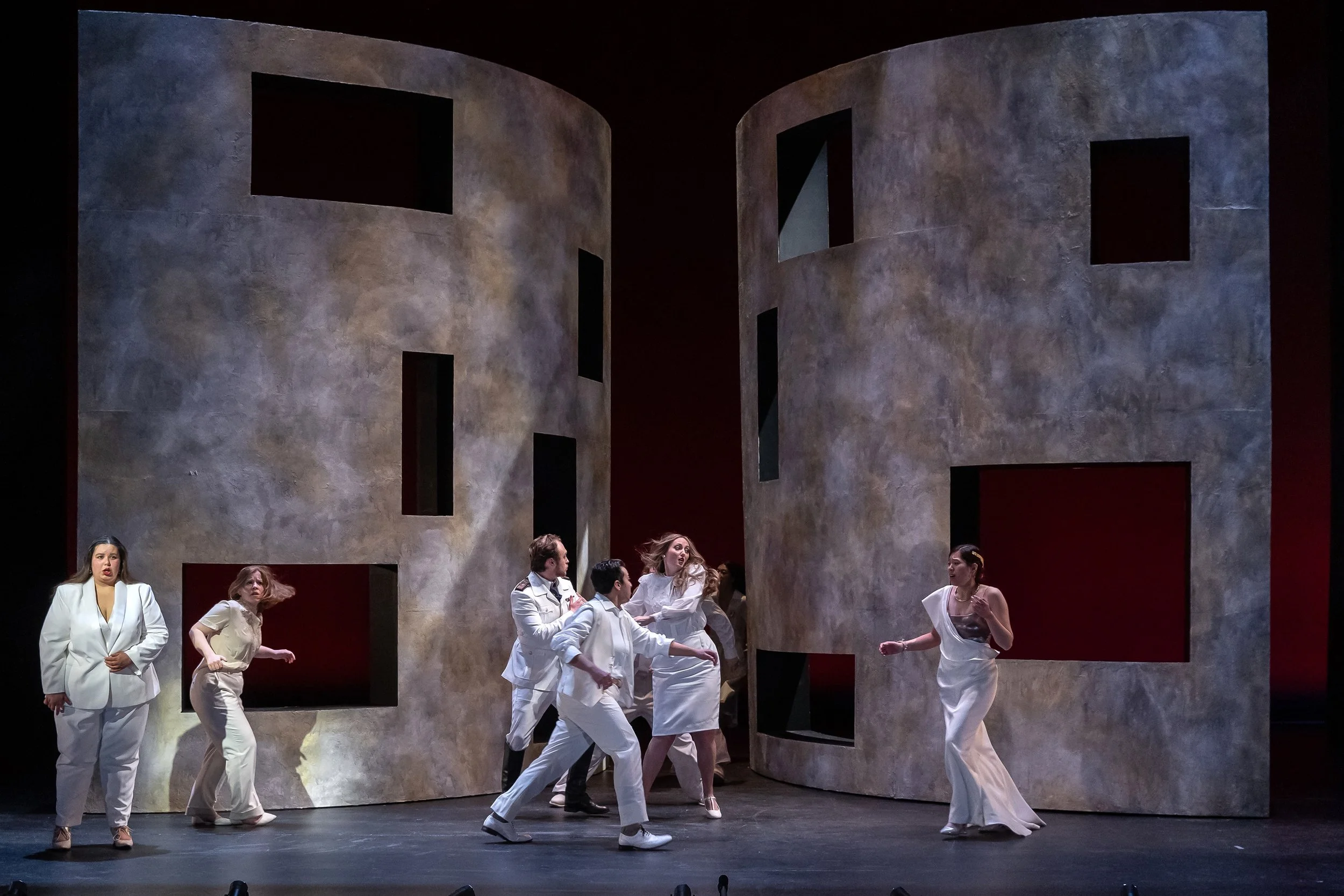 Stage performance with multiple actors in white costumes, set against a large abstract concrete-like backdrop with window cutouts, dim lighting.