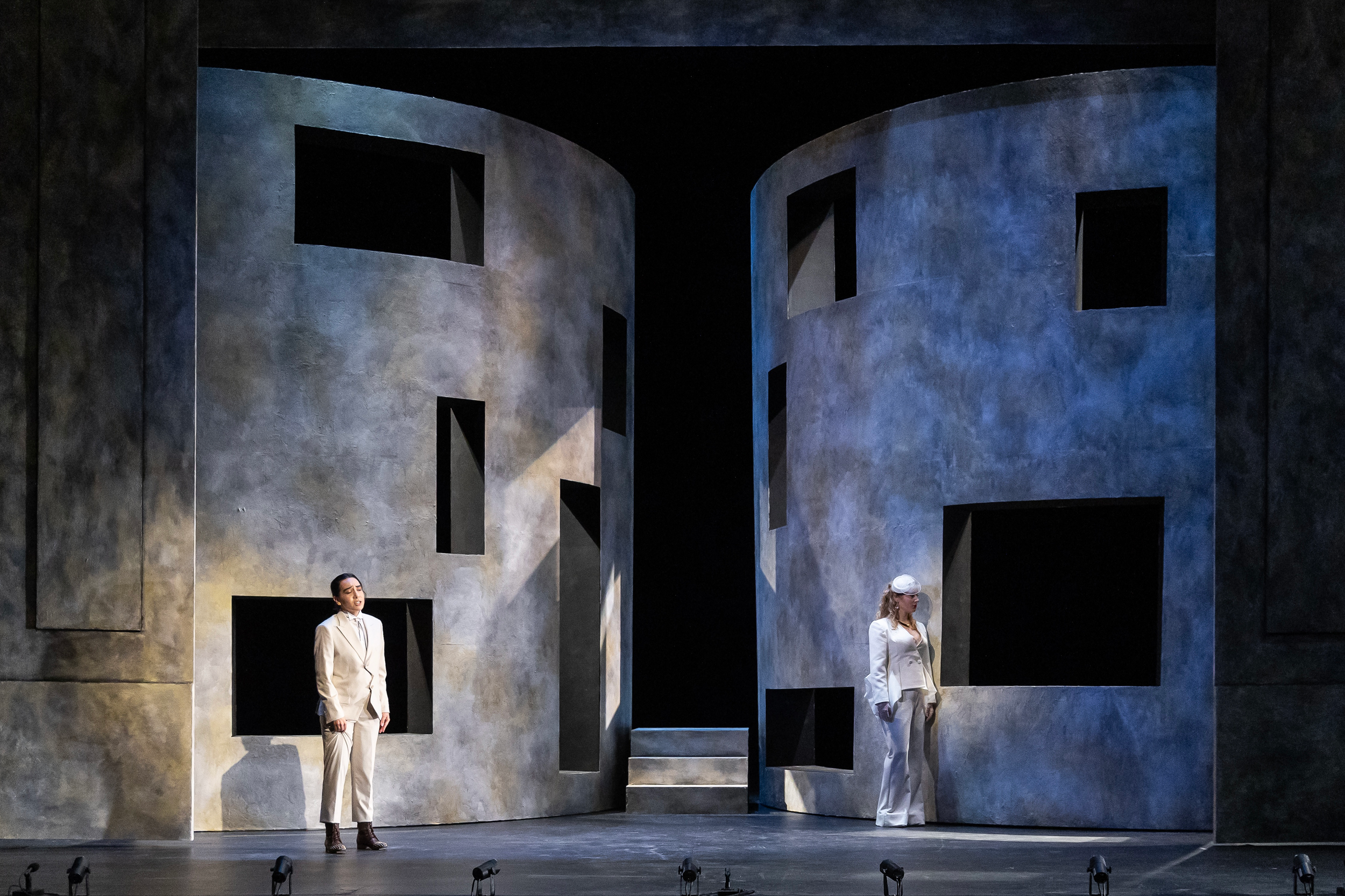 A theatrical stage with a minimalist set featuring curved walls and rectangular openings, with two women dressed in white suits standing apart.