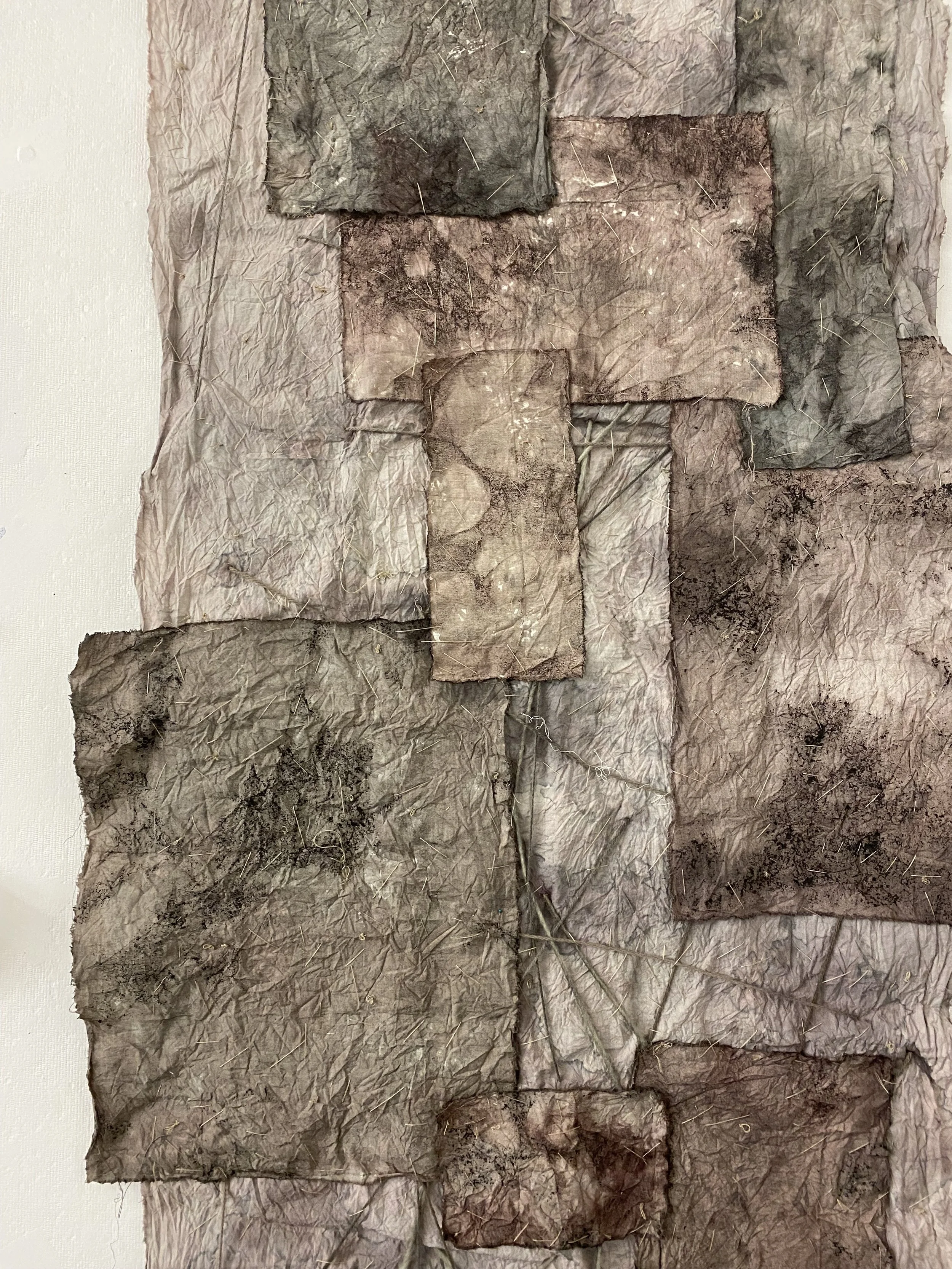Close-up view of an abstract art piece composed of crinkled, layered pieces of fabric or paper in various earthy tones, with some small patches of soil or dirt and scattered straw or thin fibers.