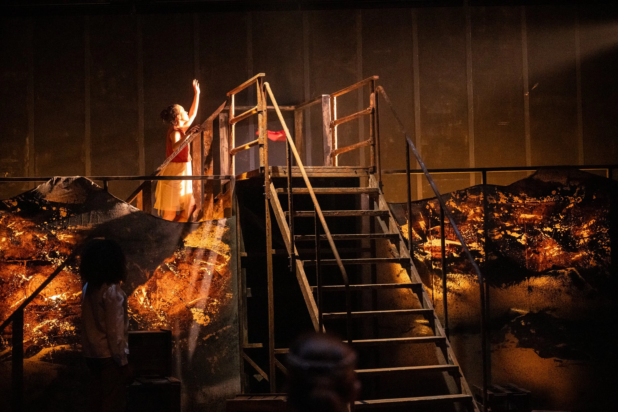 A girl in a costume with a red top and yellow skirt stands on a set of stairs and raises one arm. She is on a theatrical stage with dark background and set design that mimics a rocky landscape, with black and gold textured surfaces. There are lights 