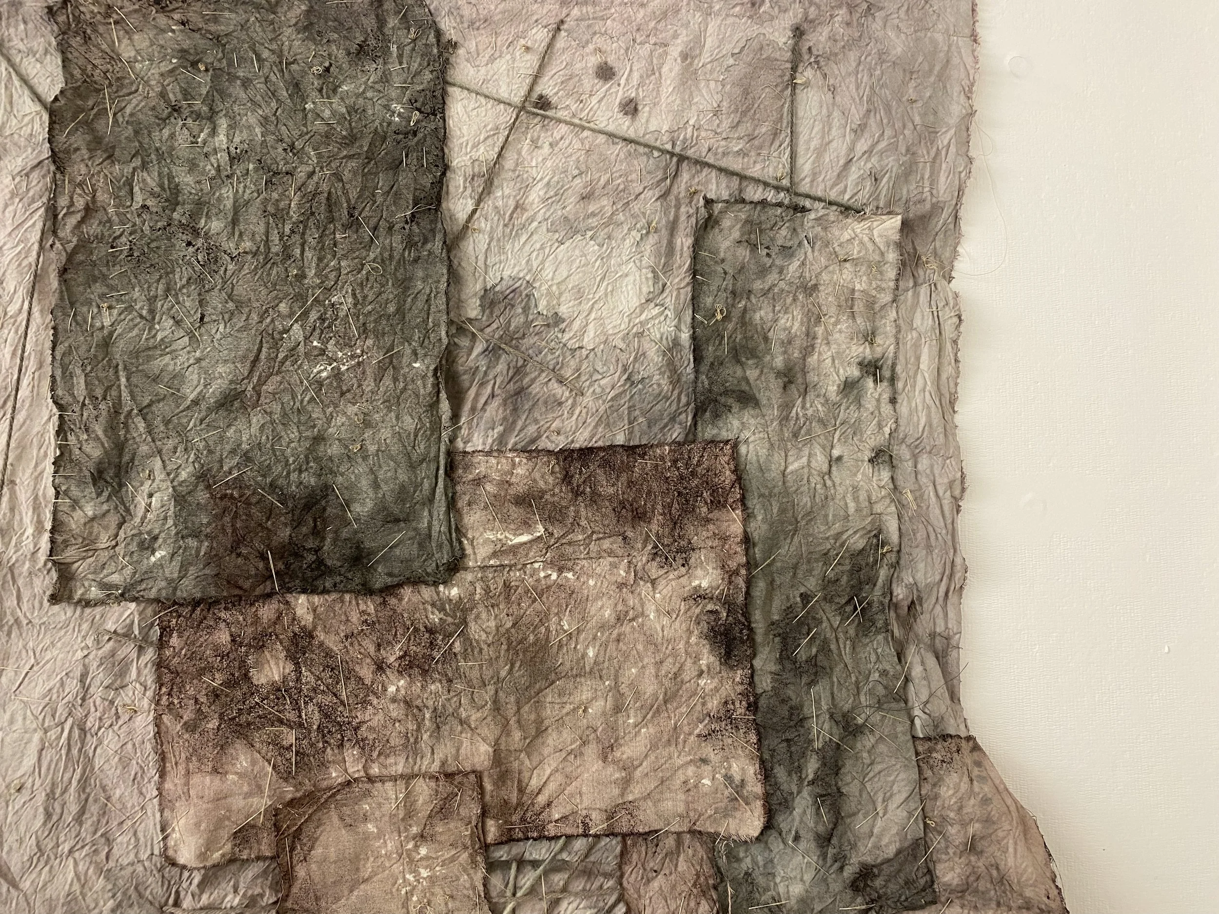 Close-up of crumpled, wrinkled fabric with embedded nails or pins and darker patches, mounted on a wall.