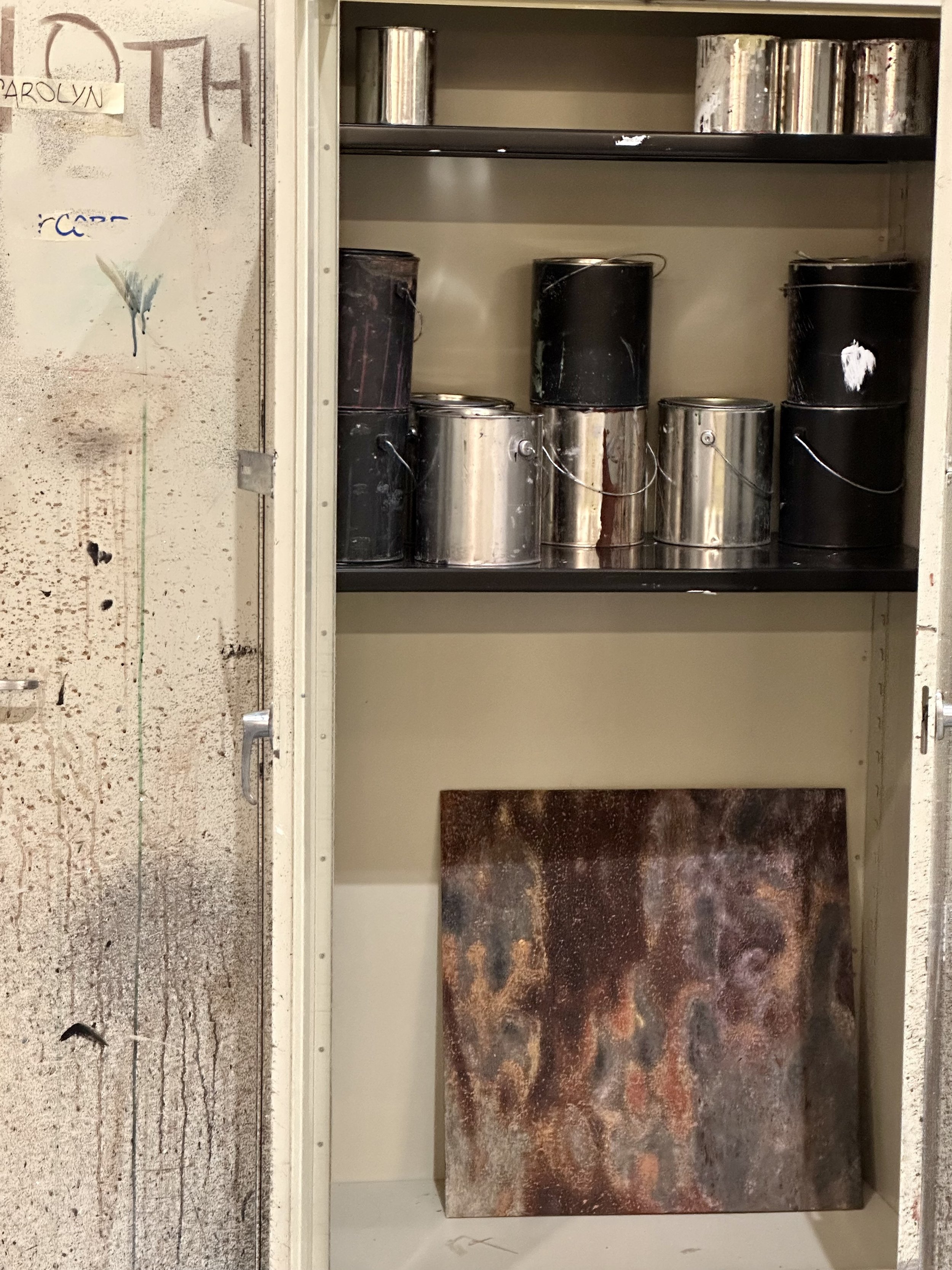 A metal shelf with several paint cans on it, surrounded by a wall with a painted and scribbled surface. Some cans are black, and some are metallic silver.