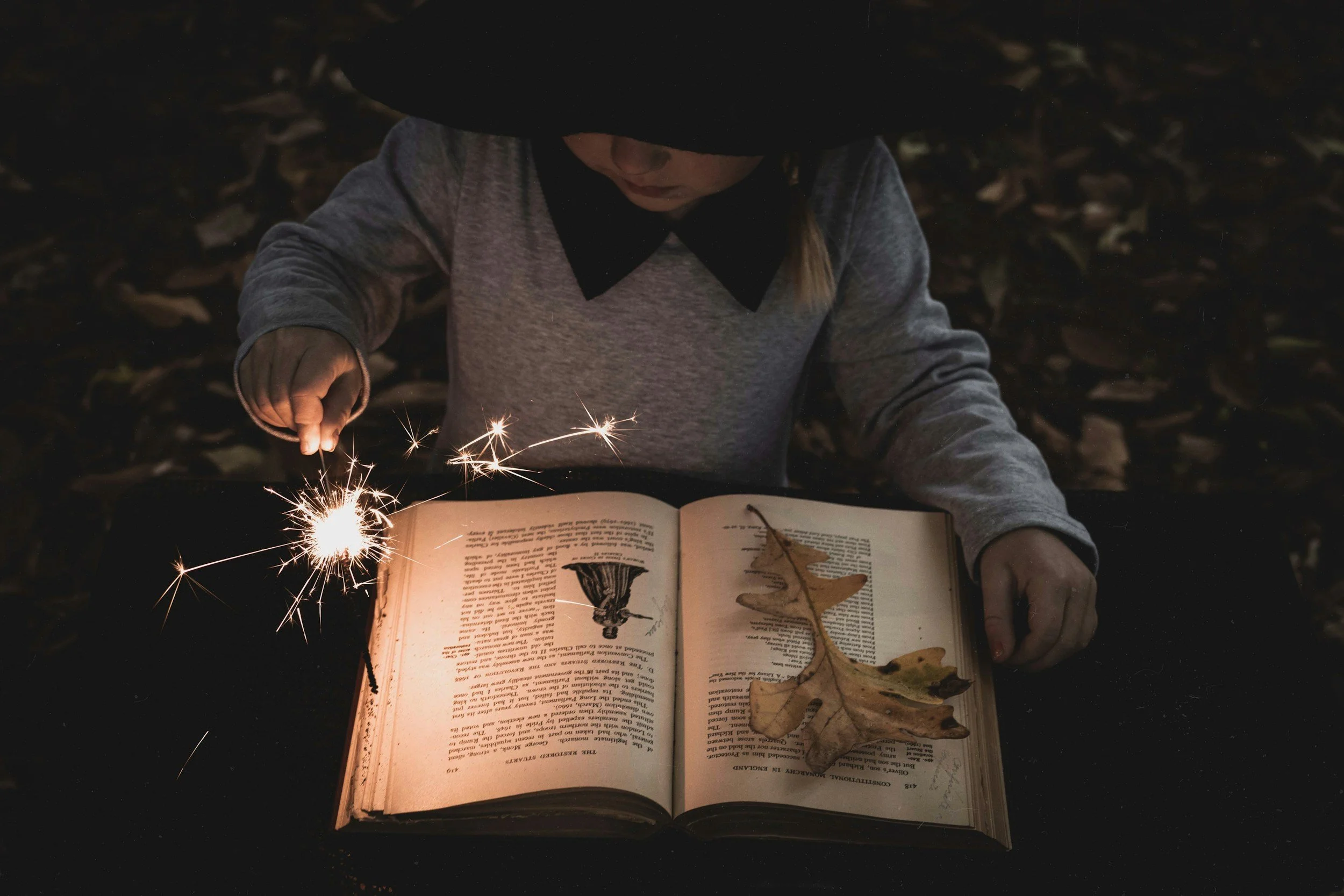 A person wearing a black hat and gray clothing is outside at night, holding a lit sparkler and reading a book on a table. The book contains illustrations of mushrooms and dried leaves are on the open pages.