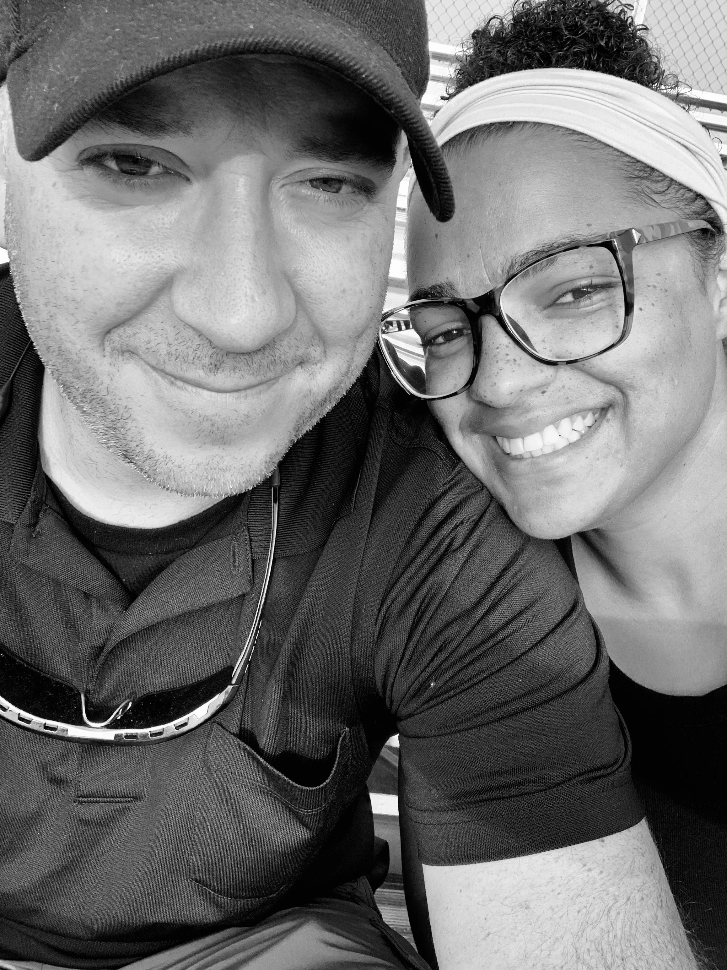 Close-up of a smiling man with glasses and a woman smiling, both in casual clothing, sitting outdoors near a chain-link fence.