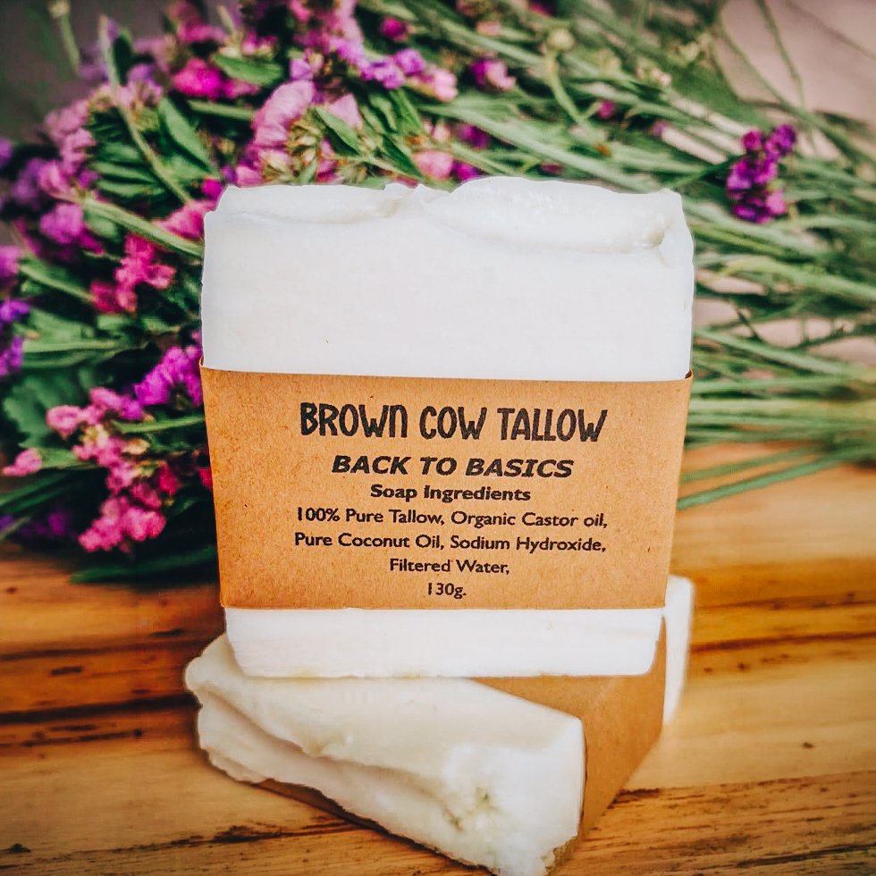 A bar of white soap labeled 'Brown Cow Tallow' with ingredients including tallow, castor oil, coconut oil, and water, placed on a wooden surface with pink flowers in the background.