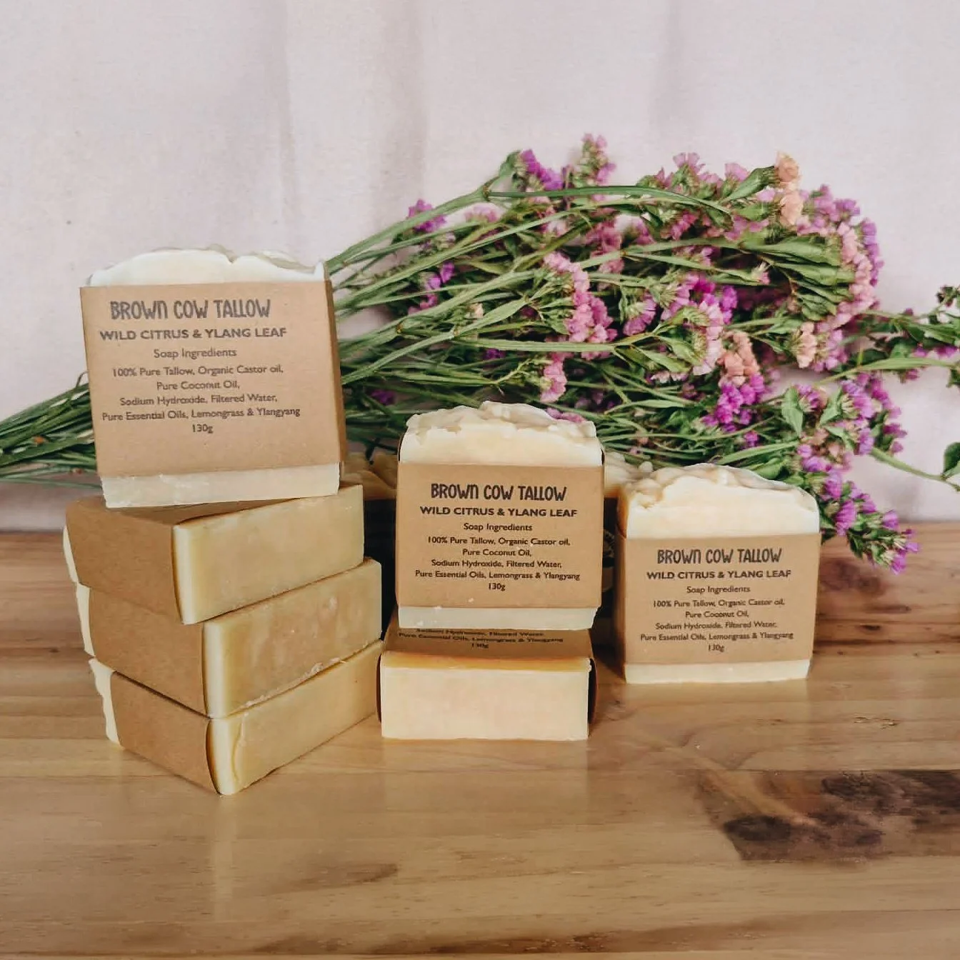 Natural soap bars with labels reading "Brown Cow Tallow, Wild Citrus & Ylang Leaf," arranged on a wooden surface with pink and purple flowers in the background.