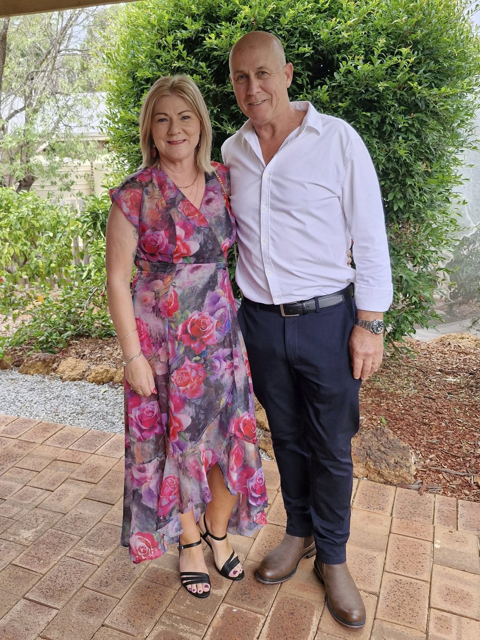 A woman in a floral pink and purple dress with black strappy heels standing next to a man in a white shirt, dark pants, and brown boots, outdoors near greenery and a wooden fence.