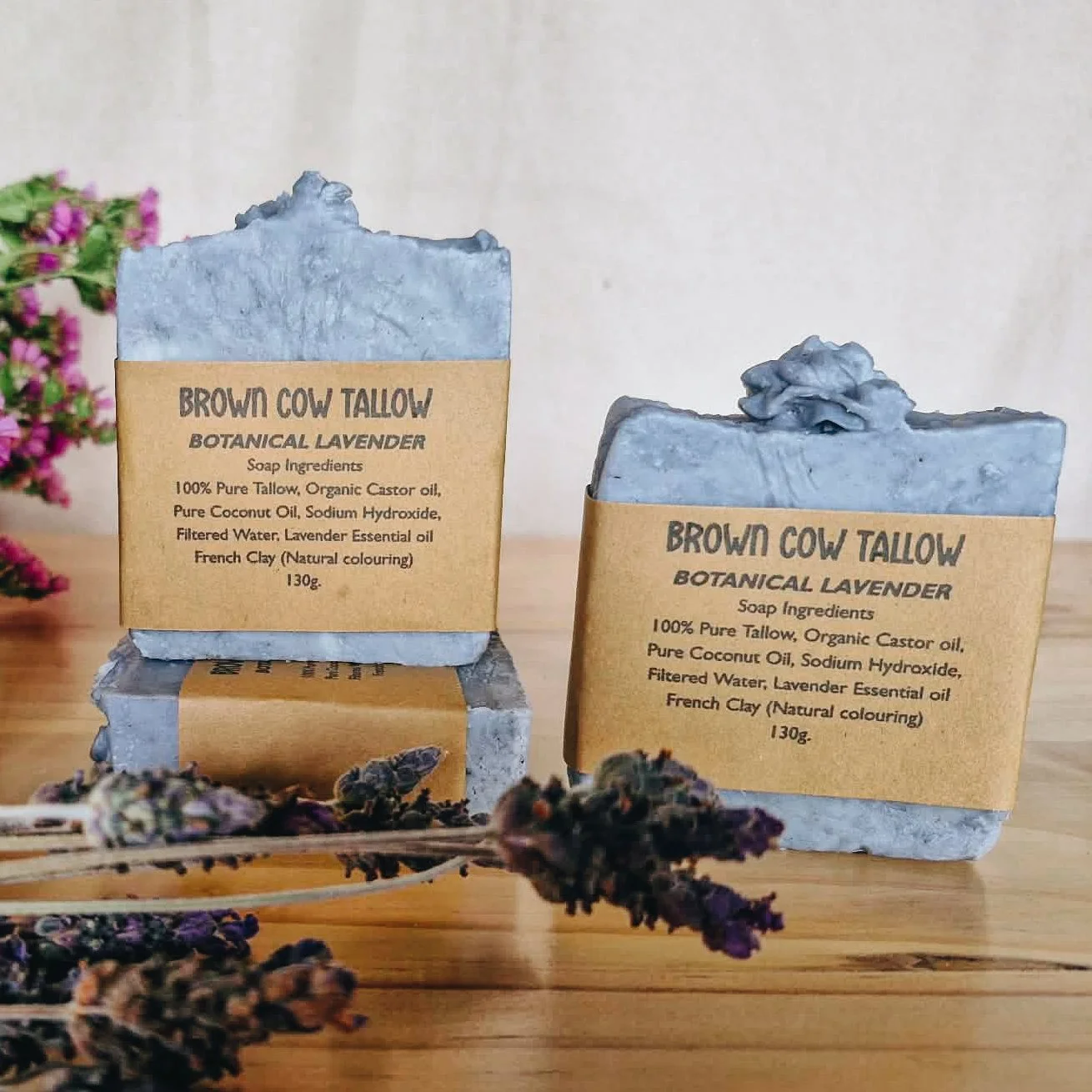 Two bars of botanical lavender soap labeled 'Brown Cow Tallow,' with natural coloring and ingredients including pure tallow, organic castor oil, coconut oil, sodium hydroxide, filtered water, lavender essential oil, and French clay, placed on a woode