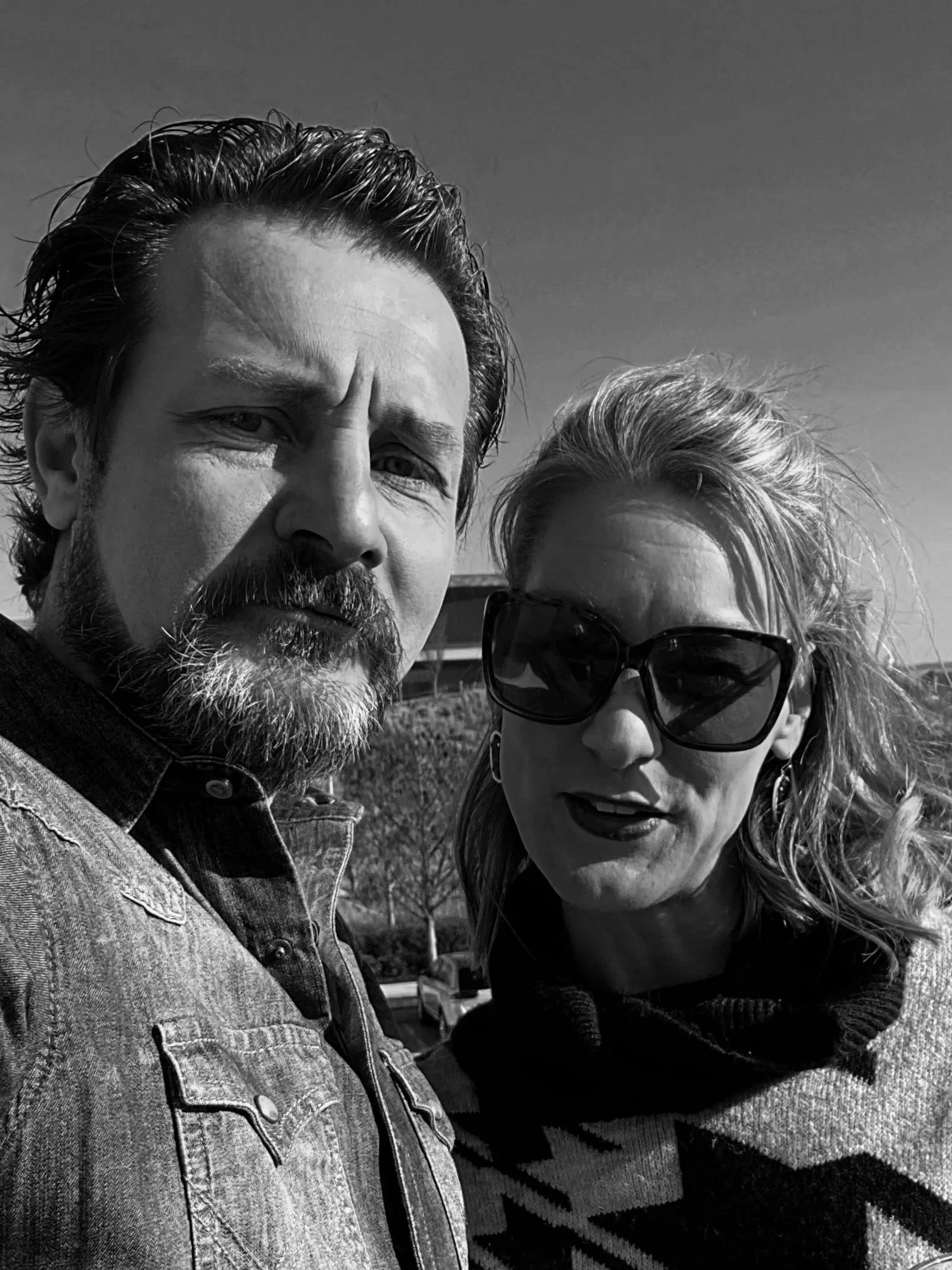 Black and white photo of a man and woman outdoors, close-up selfie, with clear sky in the background, both wearing casual clothes and sunglasses.