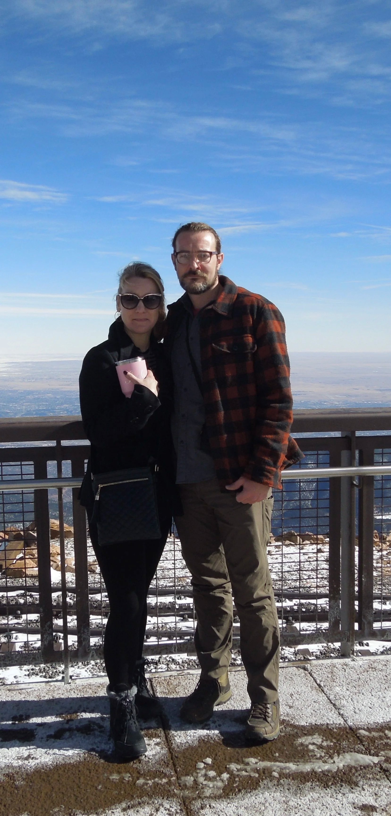 A man and woman standing on a snowy outdoor observation deck with a city skyline in the background under a blue sky.