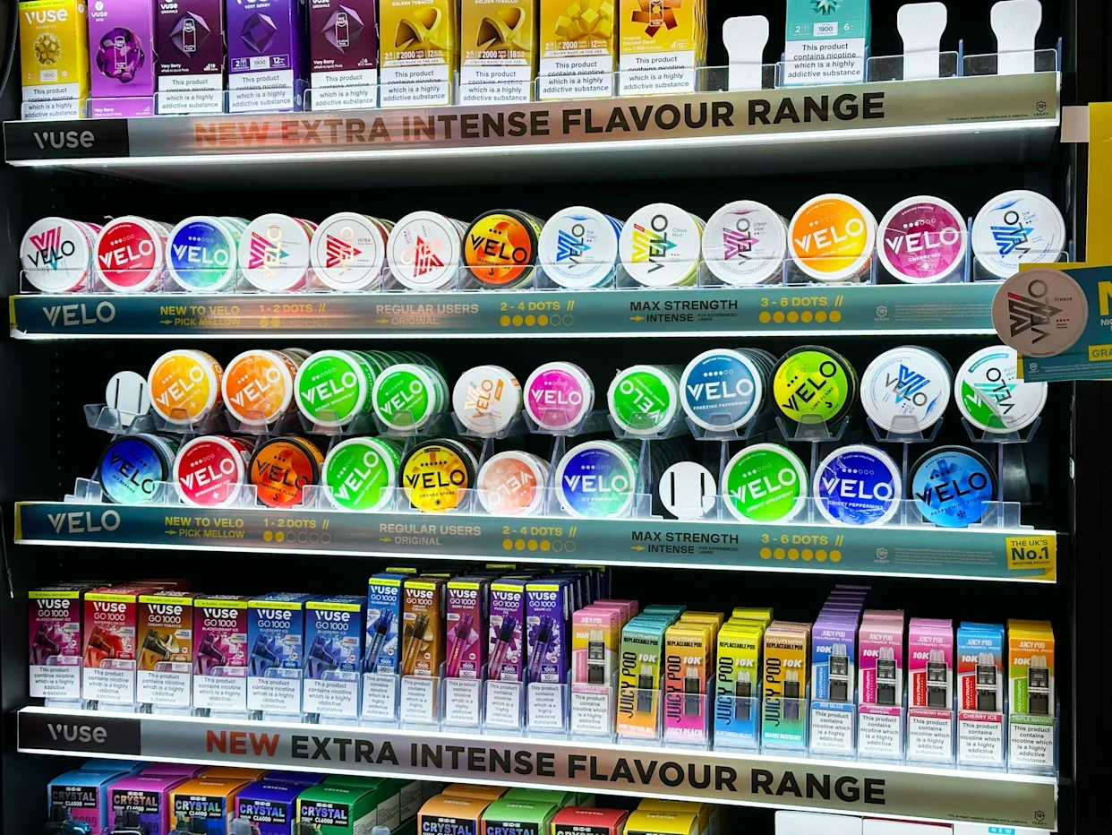 A vending shelf displaying various chewing gum and vape products, including Vuse nicotine pouches and Juicy Pod liquid vapes, arranged on multiple rows with colorful packaging.