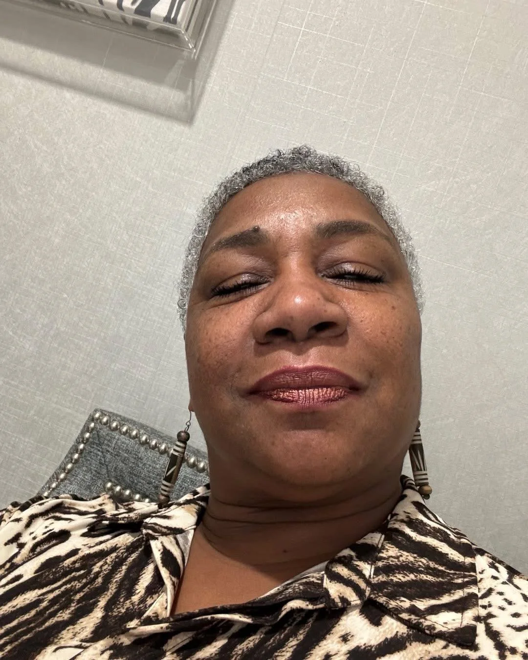 Close-up selfie of an older woman with short grey hair, wearing a patterned blouse and earrings, smiling with her eyes closed, in an indoor setting.