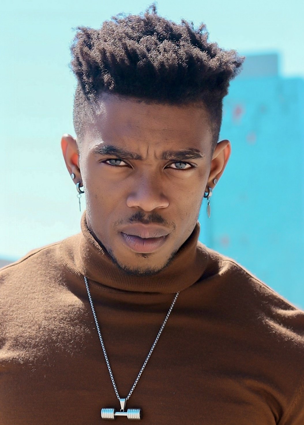 A young man with short dreadlocks looking directly at the camera, wearing a brown turtleneck and a necklace with a barbell pendant, outdoors on a sunny day.