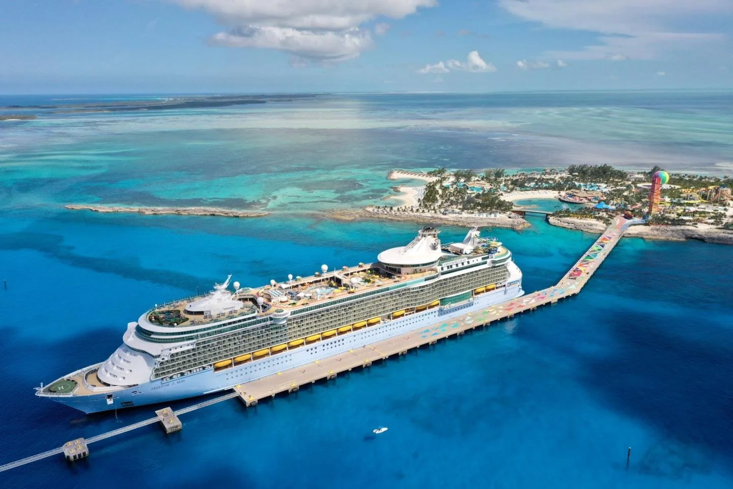 An aerial view of a large cruise ship docked at a tropical island port, with a pier connecting to a vibrant resort area with colorful water slides and lush greenery.