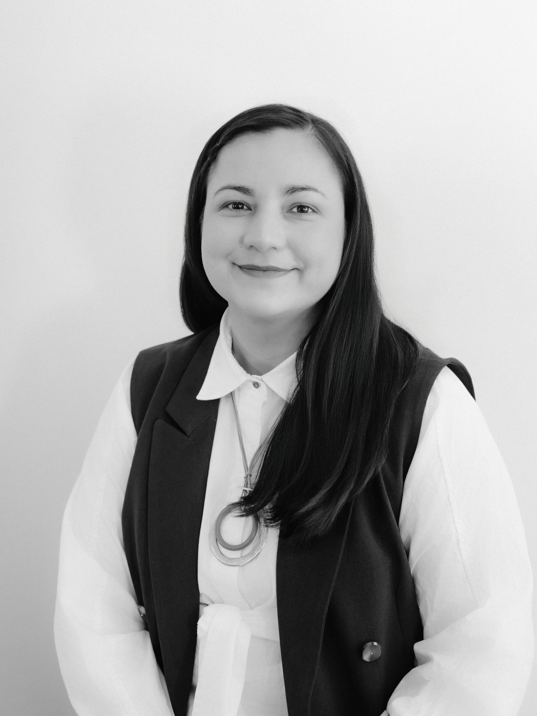 Black and white portrait of a woman with long dark hair, wearing a white shirt, black vest, and a necklace, smiling softly against a plain background.