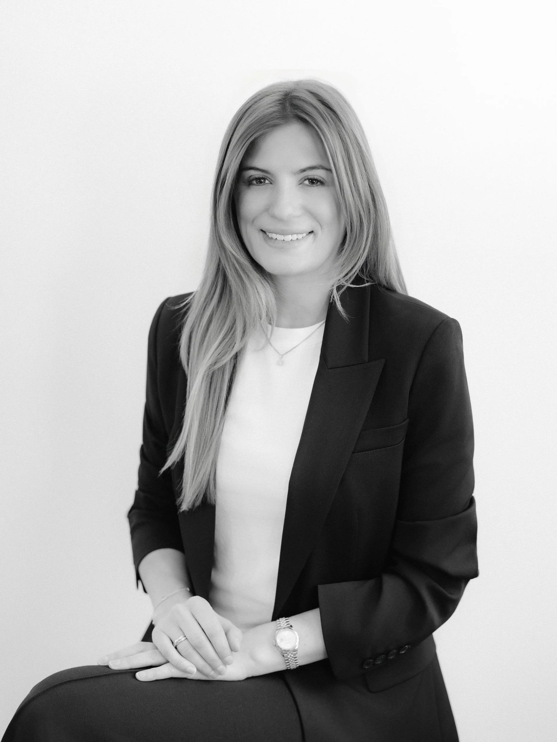 Black and white portrait of a woman with long hair wearing a blazer, white top, and jewelry, sitting with her hands resting on her knee against a plain background.