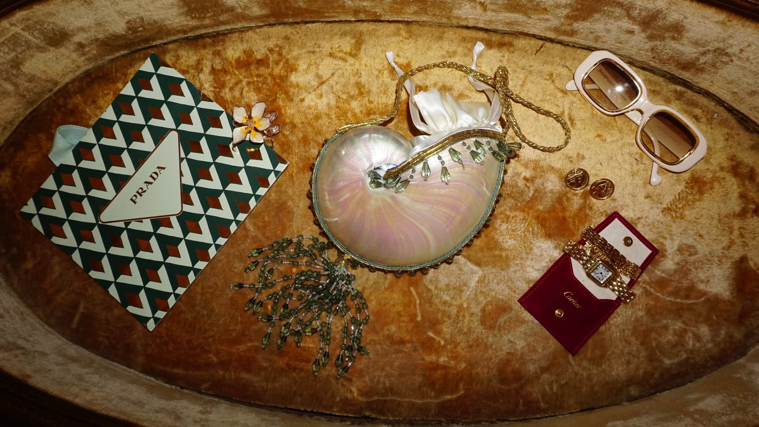 Assorted luxury accessories on a rounded wooden surface, including a Prada shopping bag, a iridescent clutch, a pair of white sunglasses, gold earrings, a floral hairpin, a pearl necklace, and a Cartier wristwatch in a red velvet box.