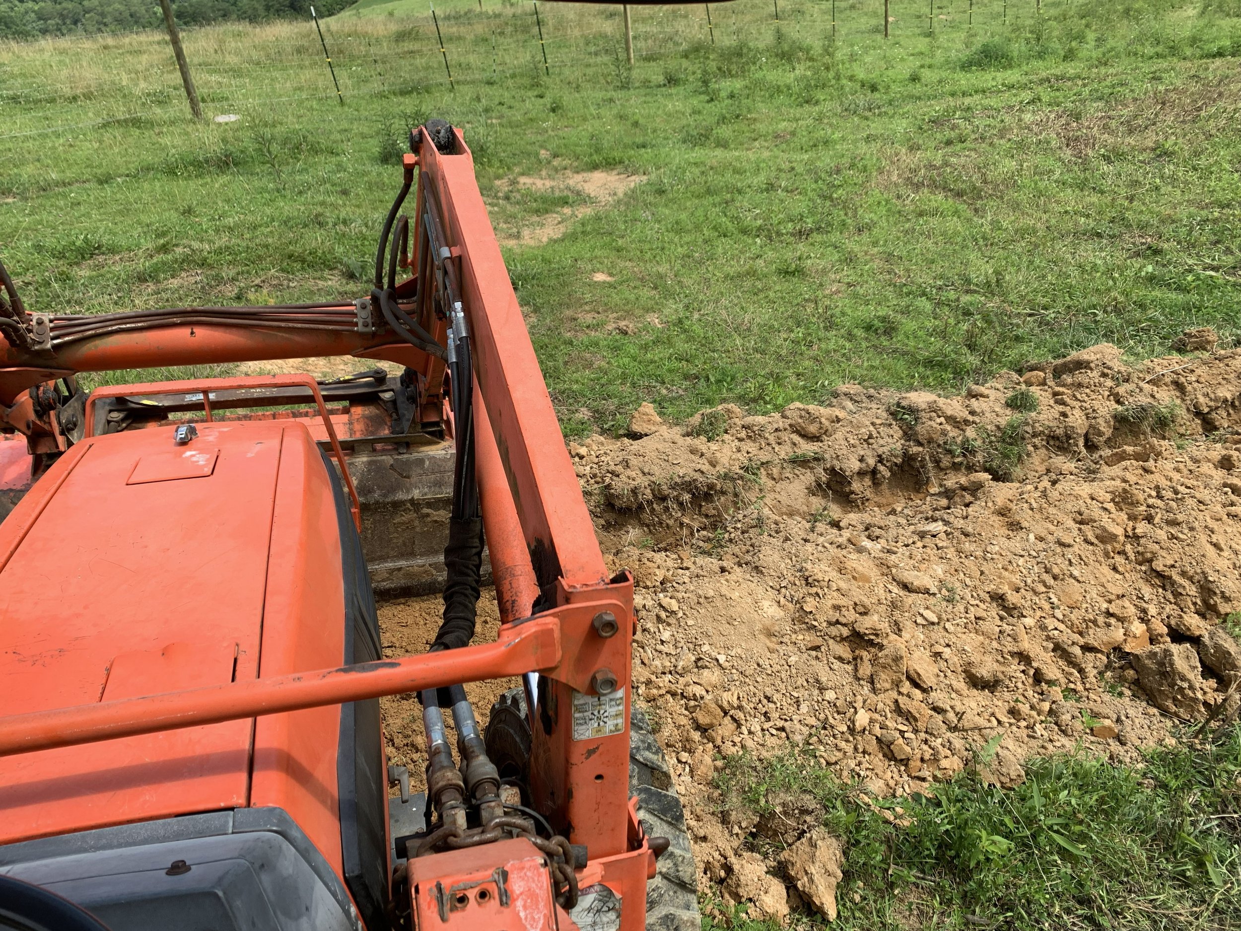 Front view of a bright orange tractor with a hydraulic arm at the construction site on grassy land, with a patch of loose dirt and a wire fence in the background.