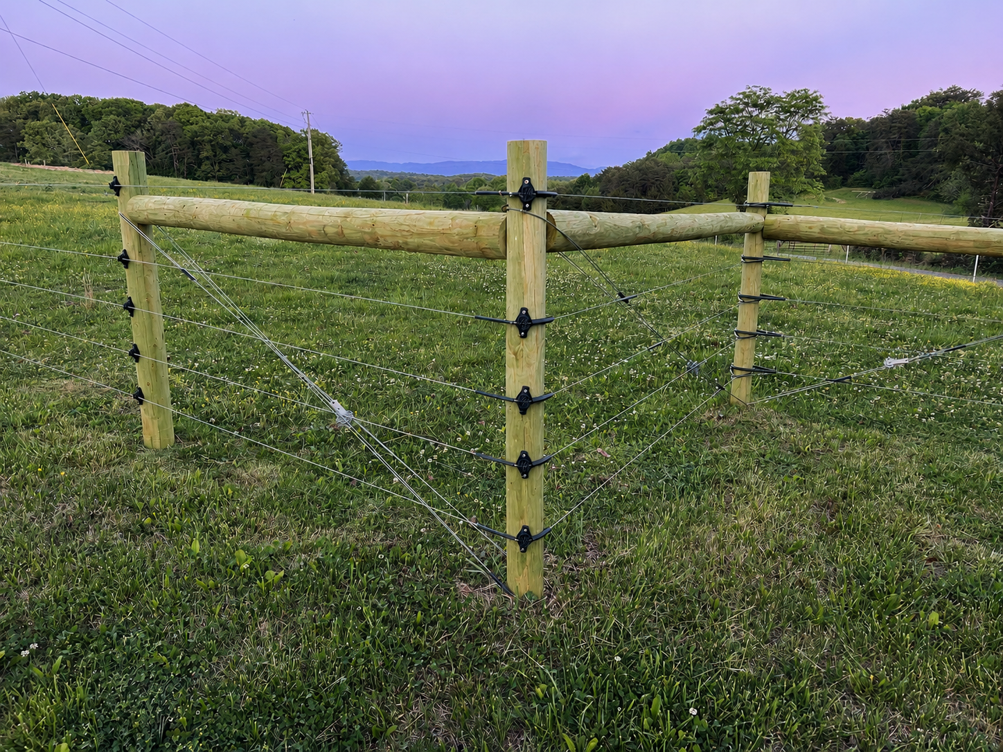 A wooden fence with wire strands stretches across a grassy field in a rural area with trees, hills, and electrical poles in the background during twilight.