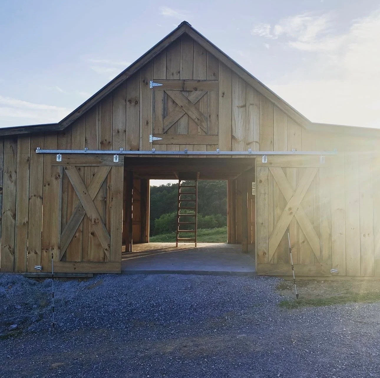 Front view of a wooden barn with sliding doors, open to show the interior and a ladder inside, set against a background of green landscape and daytime sky.