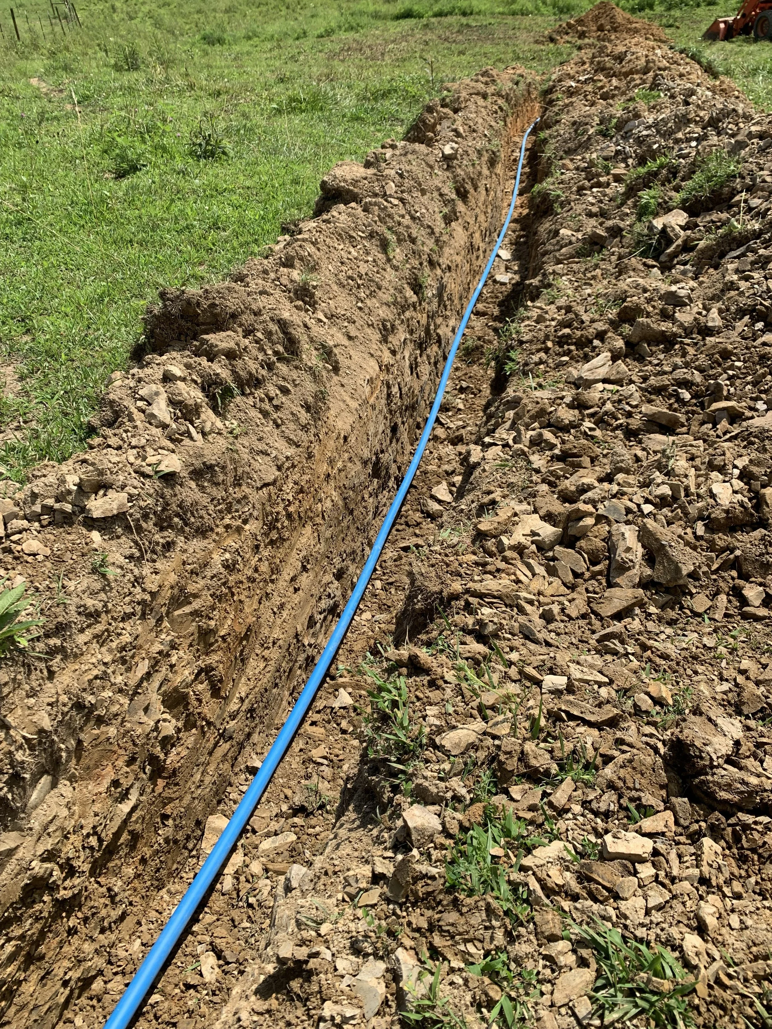 A trench in the ground with a blue pipe running through it, surrounded by dirt and small rocks, on a grassy field.
